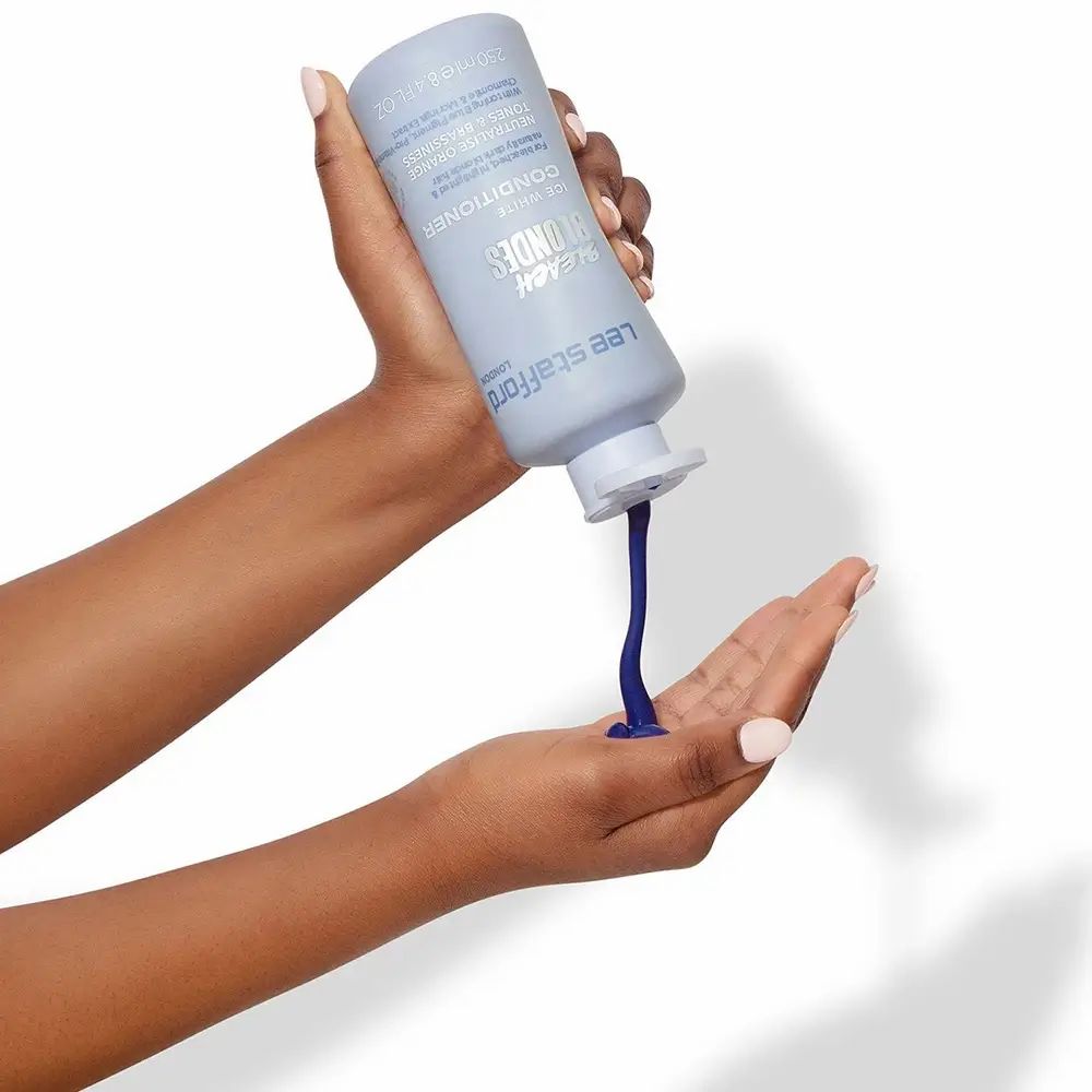 Mani che tengono un flacone azzurro chiaro. Il prodotto viene versato nel palmo. Prodotto: LEE STAFFORD Bleach Blondes Conditioner.