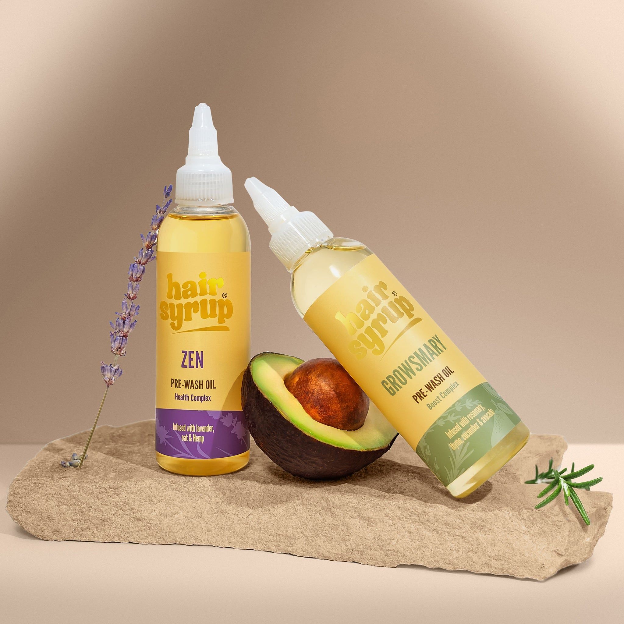 Due bottiglie di liquido giallo con etichette. Una bottiglia appoggiata a un avocado. Lavanda e pietra sullo sfondo.