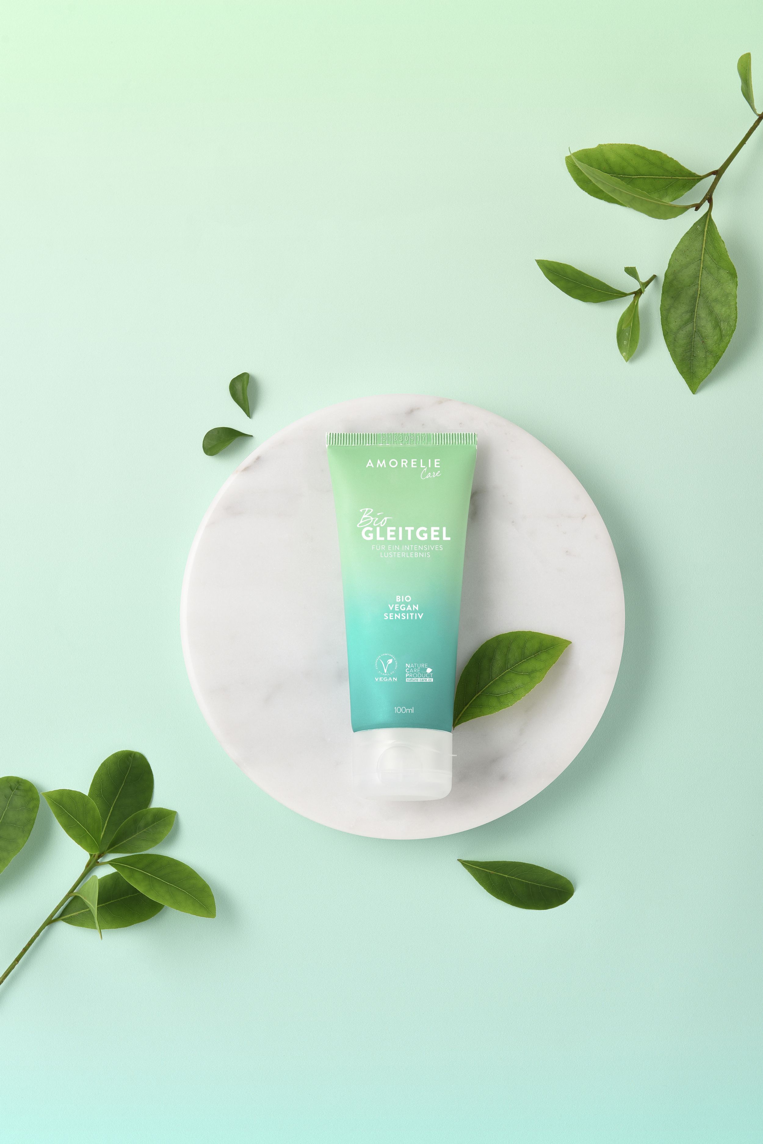 Tubo verde su piatto bianco, circondato da foglie. Scritta: AMORELIE Care, Bio Gleitgel, Bio Vegan Sensitiv. Sigillo vegano.