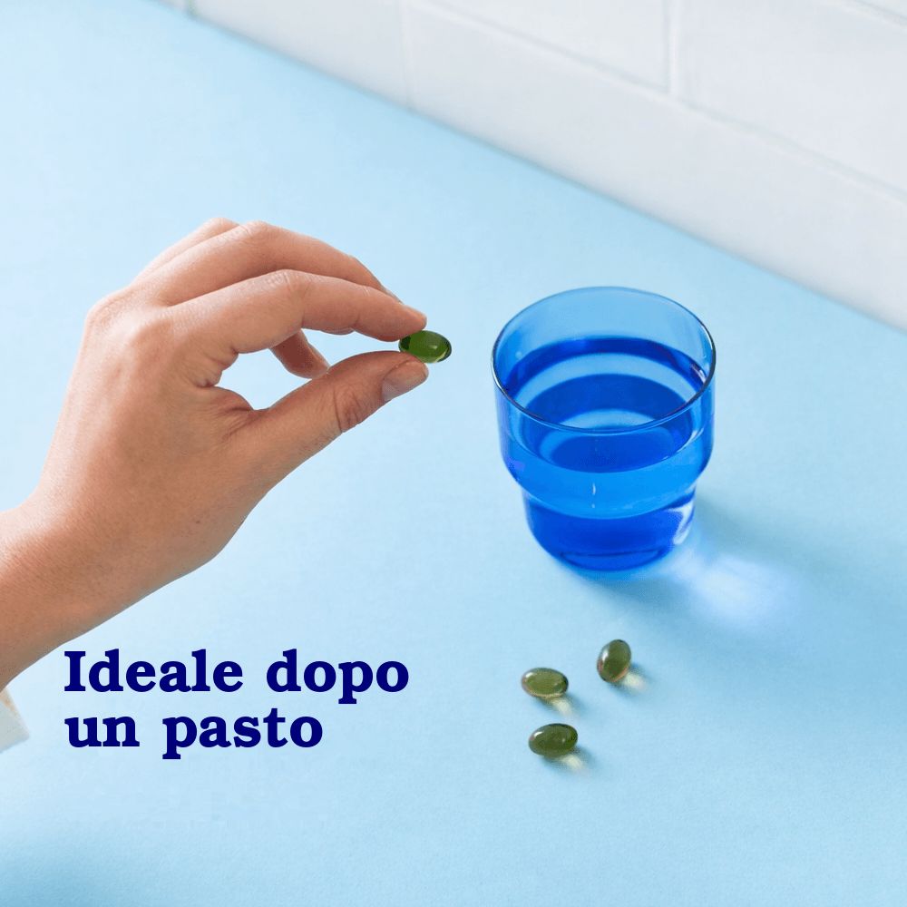 Una mano tiene una capsula verde accanto a un bicchiere blu d'acqua e altre tre capsule.