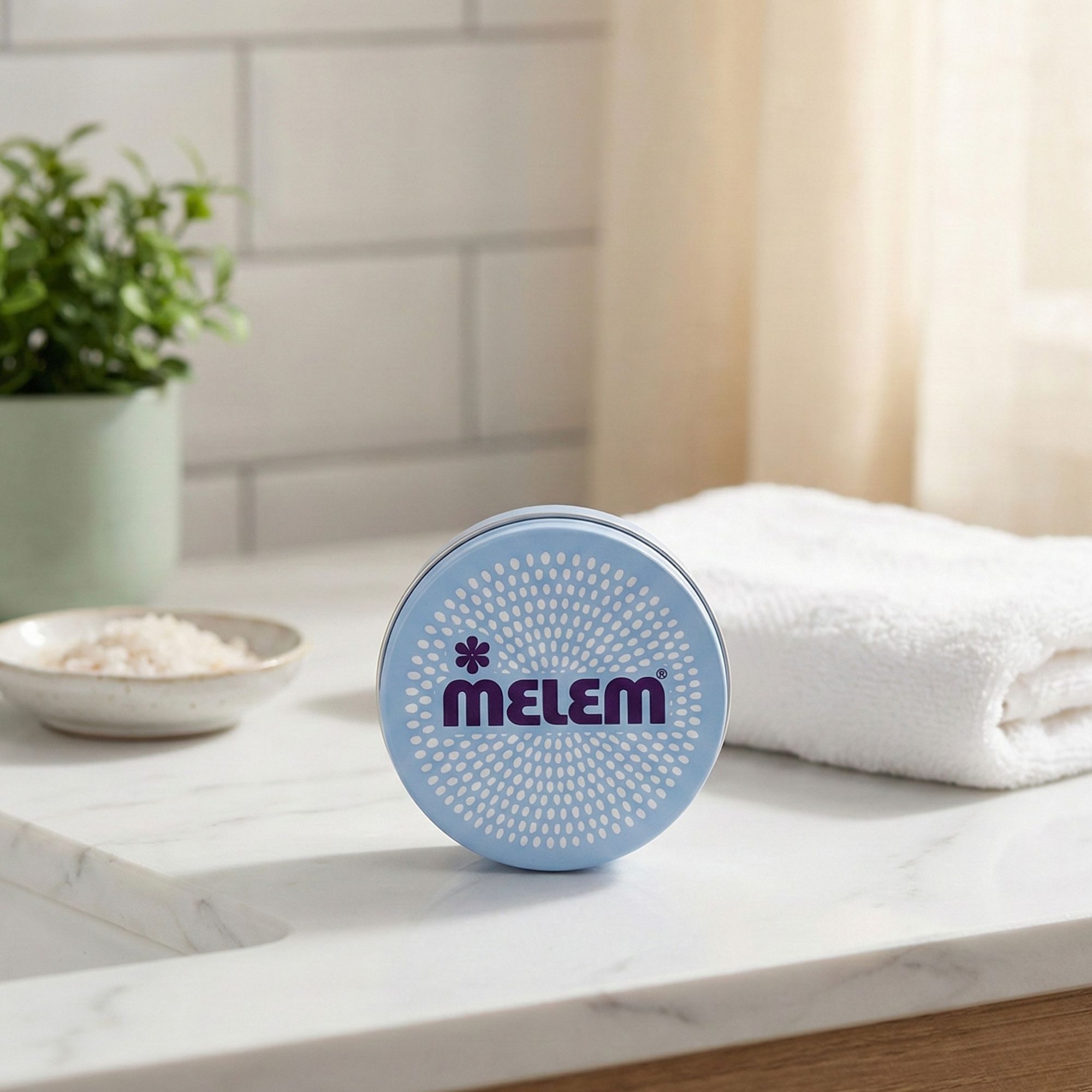 Prodotto rotondo blu chiaro con scritta viola "MELEM". Su una superficie bianca, accanto a un asciugamano e una pianta.