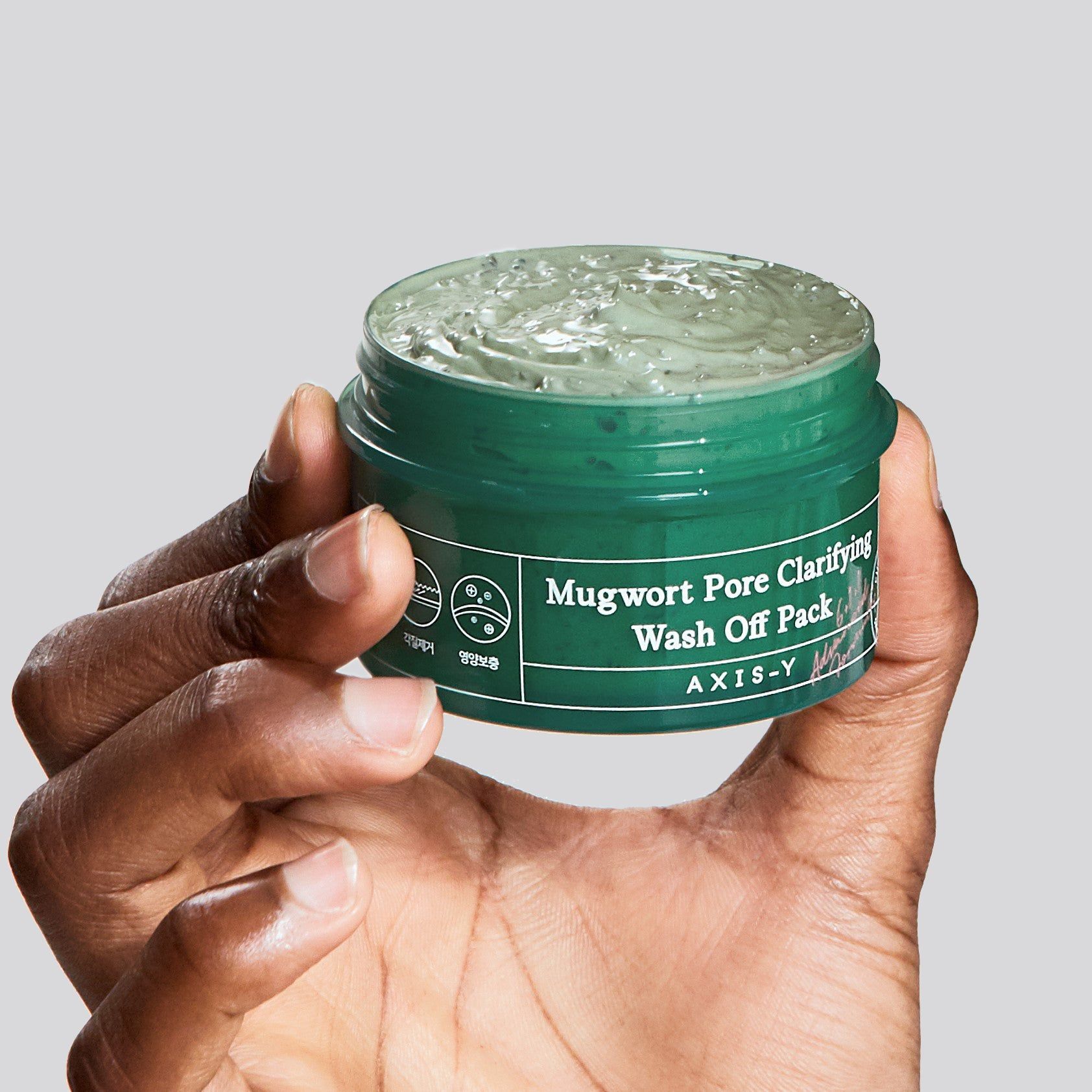 Mano che tiene un vasetto verde di maschera per il viso. Scritta: Mugwort Pore Clarifying Wash Off Pack. Marchio: AXIS-Y.