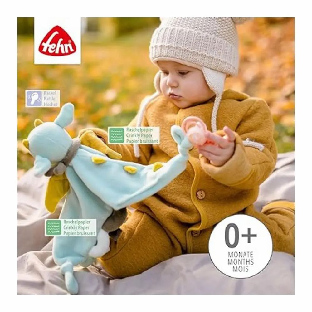 Bambino tiene in mano un peluche a forma di drago blu e giallo. Testo: 0+ mesi. Marchio: fehn. Testo: Carta scricchiolante.
