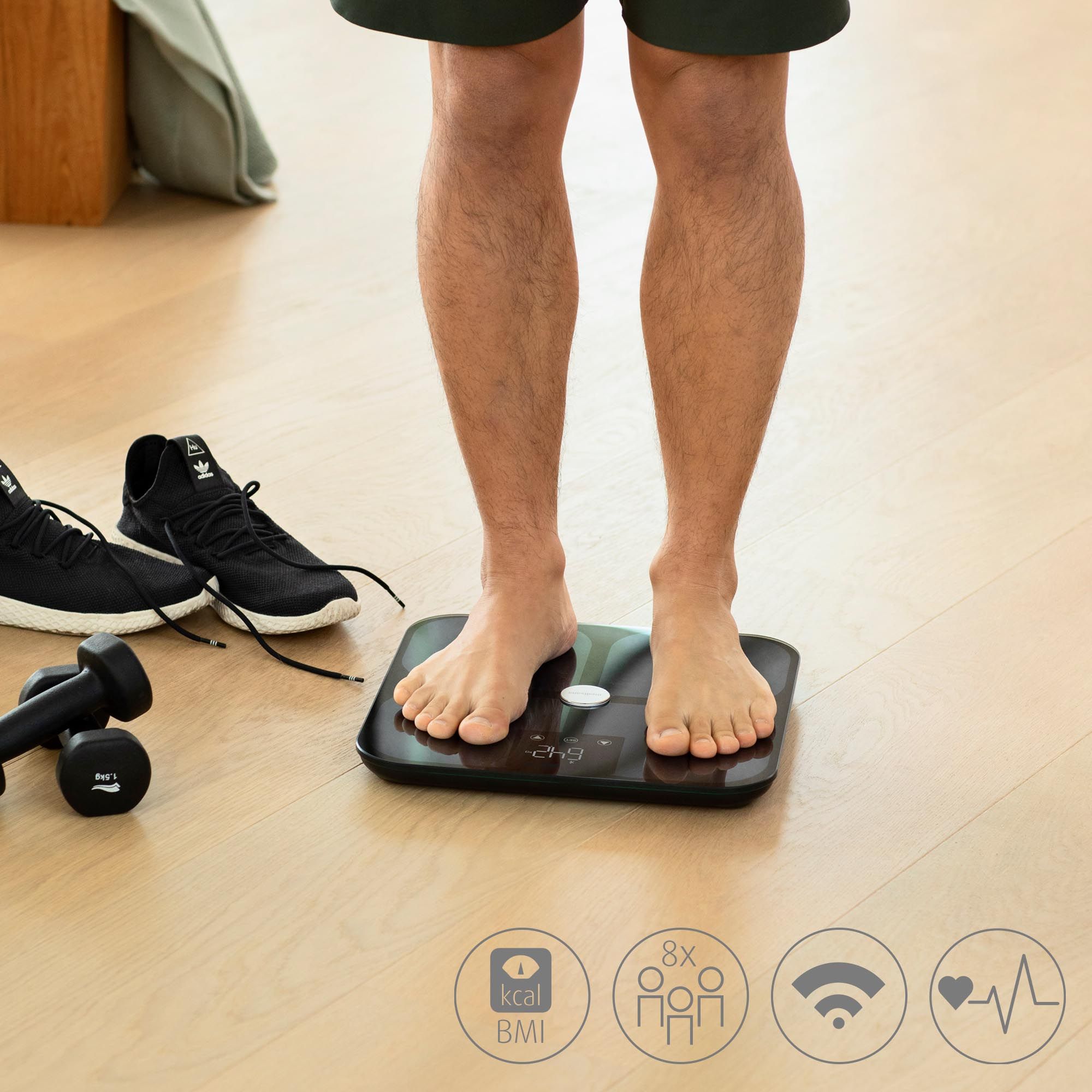 Persona in piedi a piedi nudi su una bilancia per l'analisi corporea nera. Piedi sulla bilancia, accessori sportivi accanto.