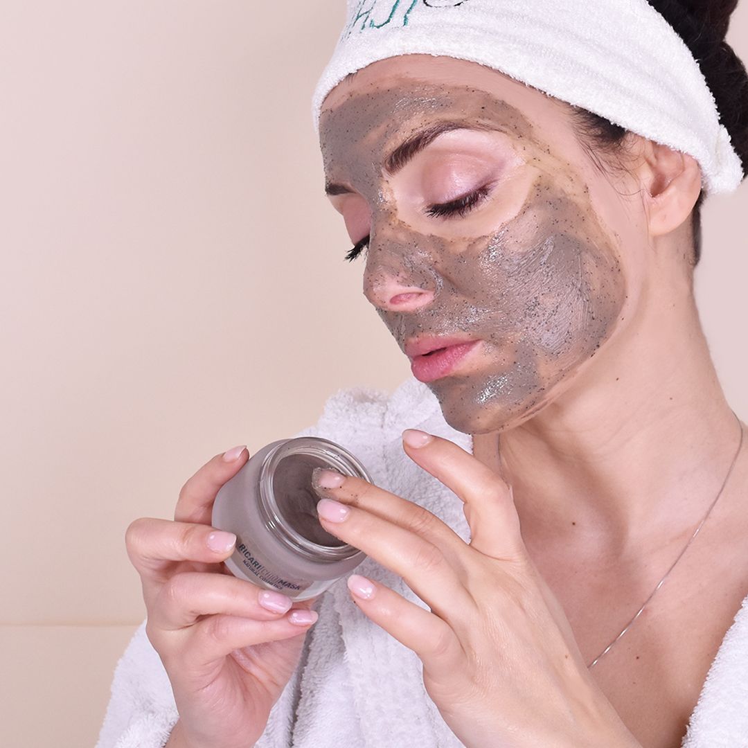 Donna con maschera viso. Tiene un barattolo. Indossa un asciugamano e un accappatoio.