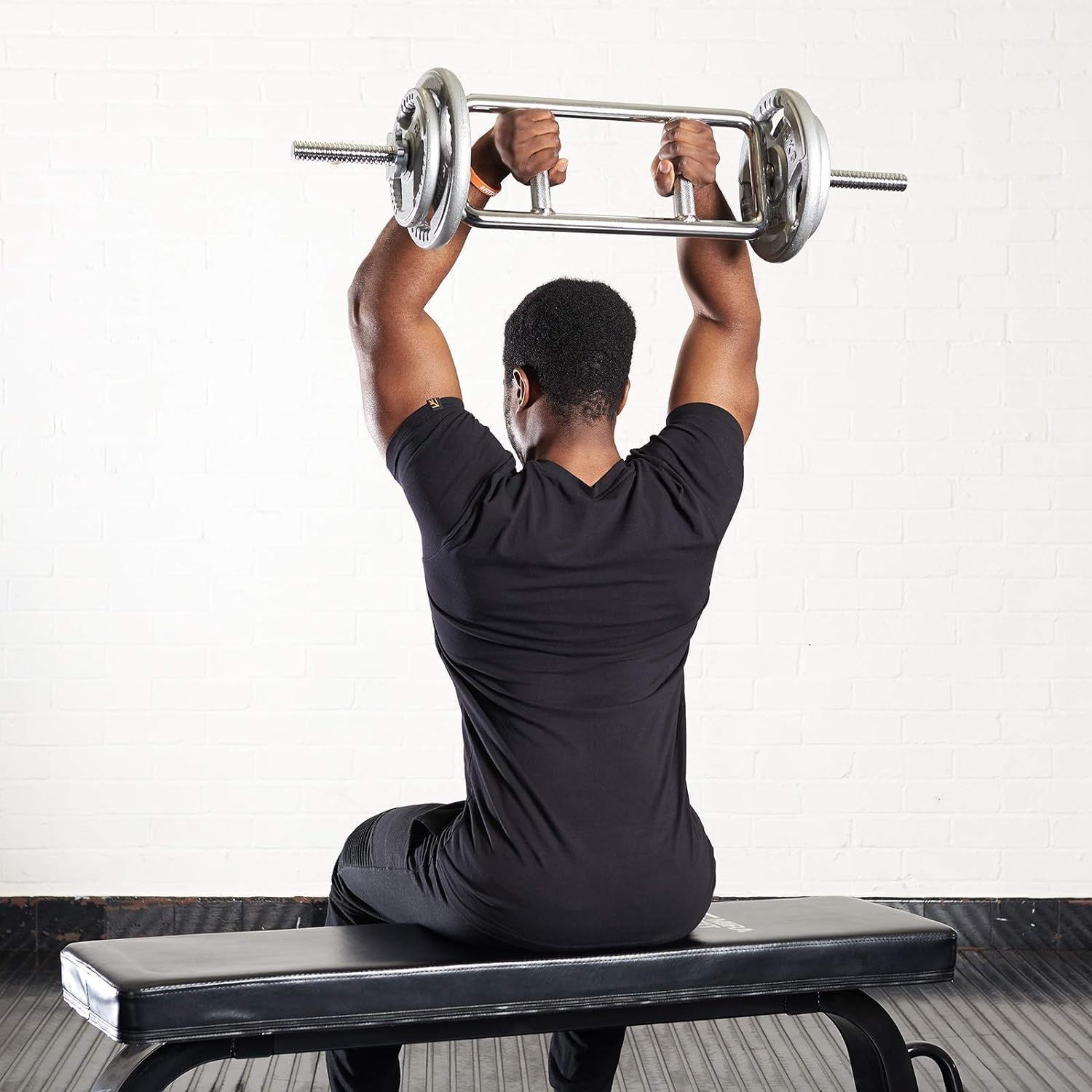 Persona che solleva una barra per tricipiti sopra la testa. Maglietta nera, seduta su una panca nera davanti a un muro bianco.