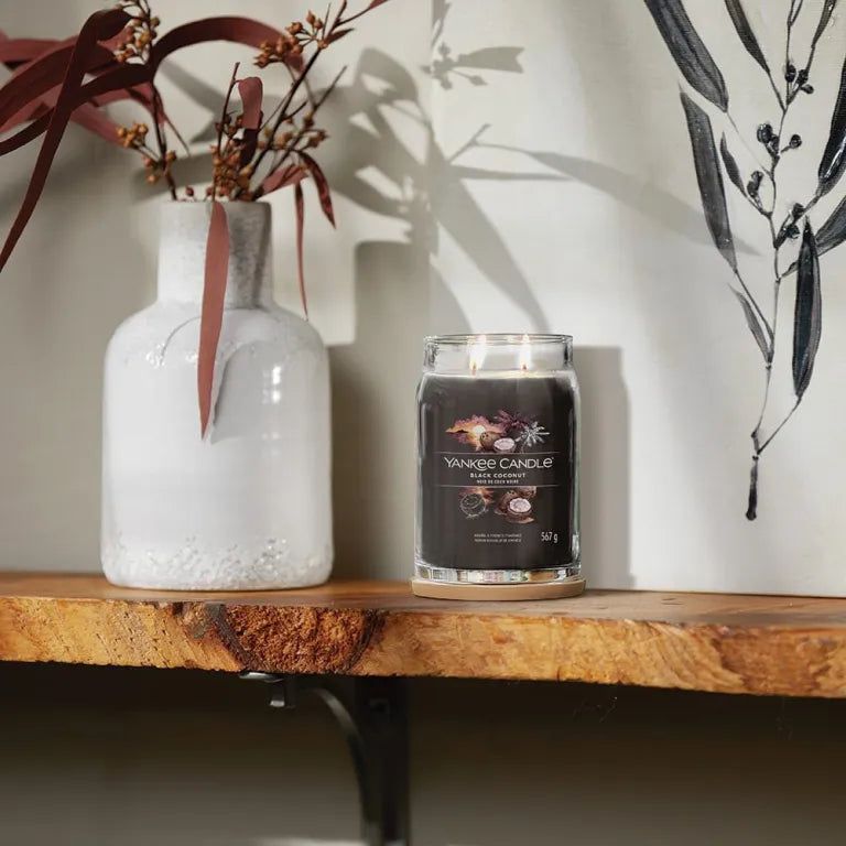 Candela grigio scuro su mensola in legno. Accanto a vaso con rami. Opera d'arte sullo sfondo. Candela con etichetta.