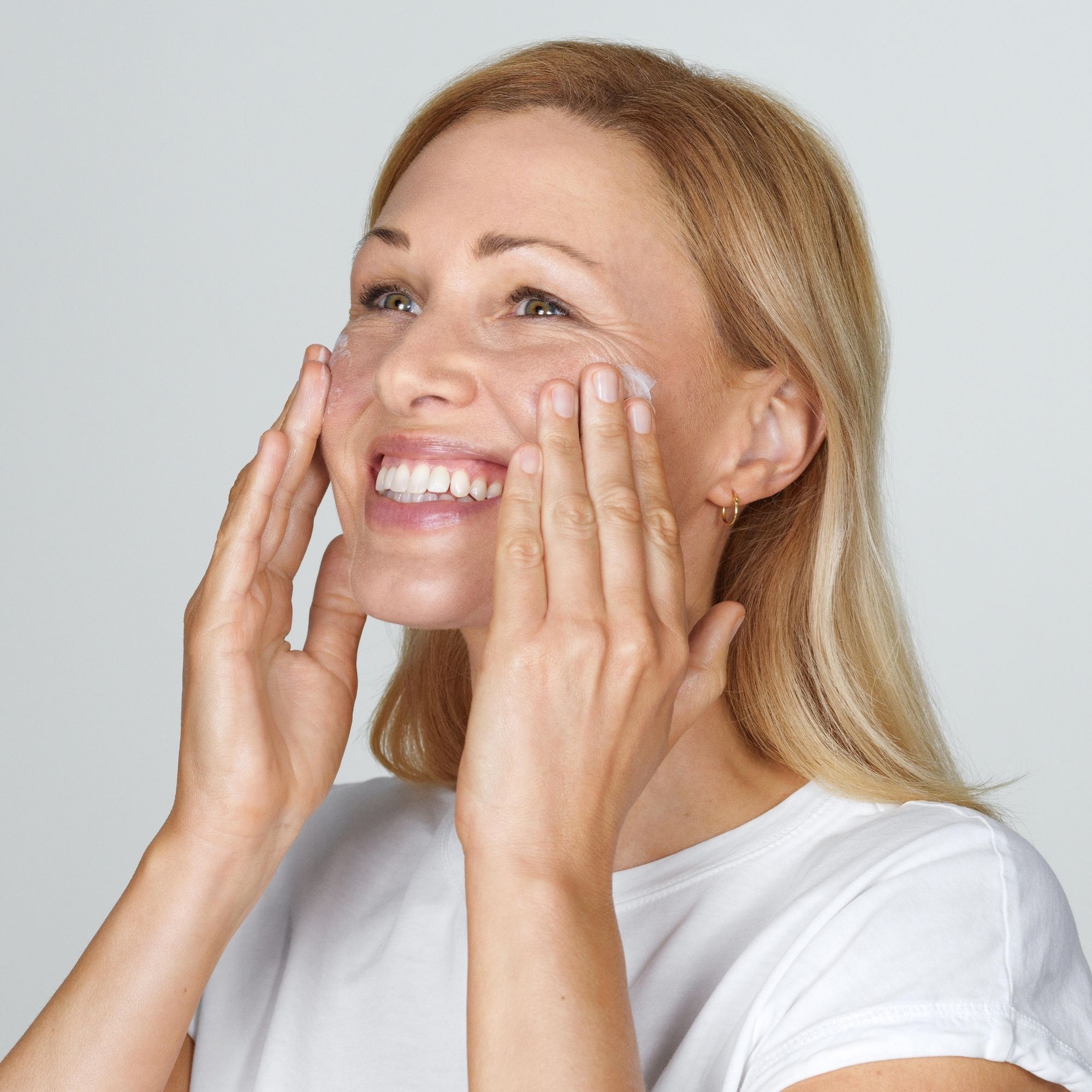 Donna applica crema sul viso. Sorride e guarda in alto. Pelle chiara, capelli biondi, maglietta bianca.