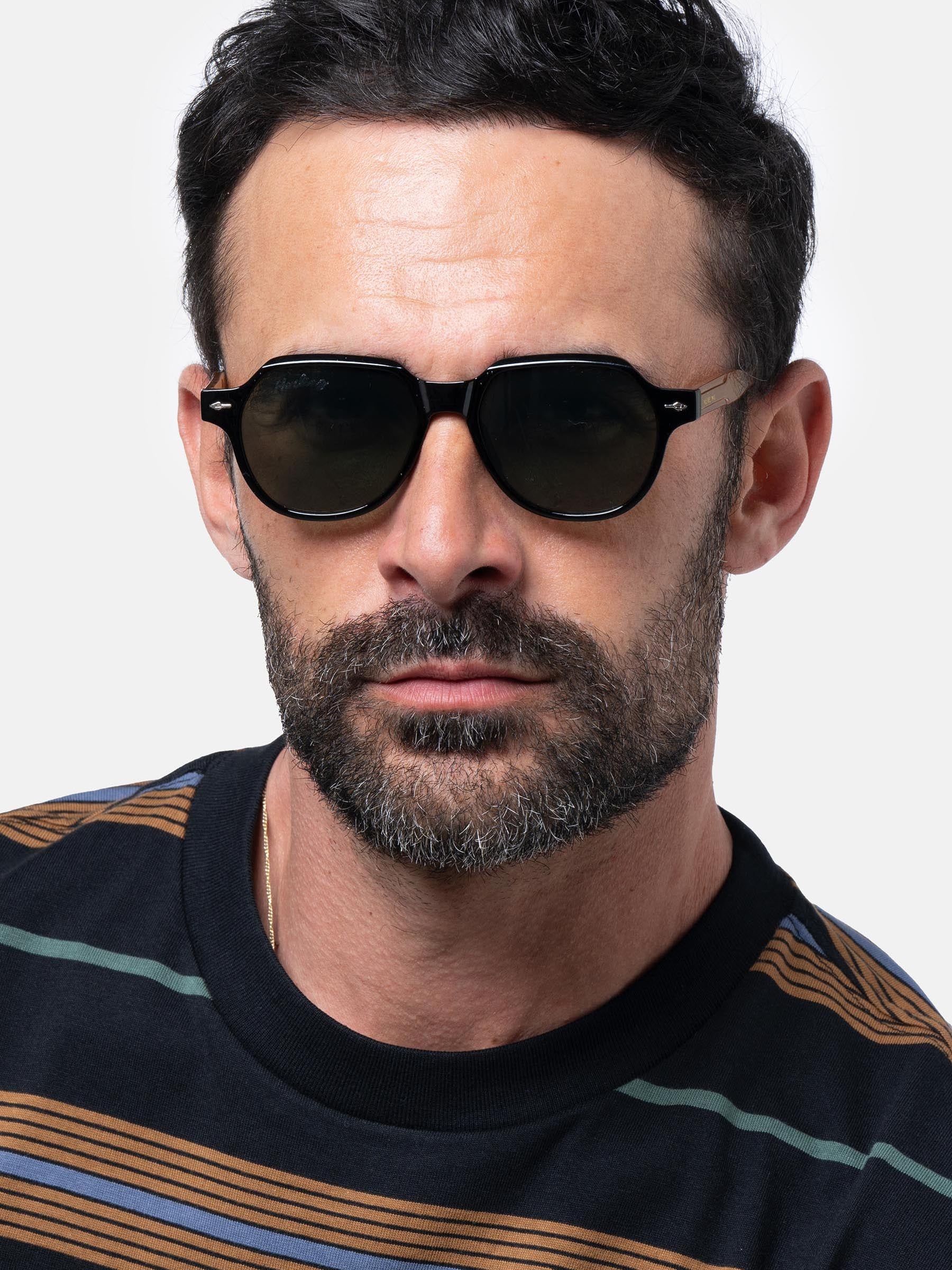 Uomo con occhiali da sole neri. Forma angolare, lenti verdi. Logo Herling visibile.