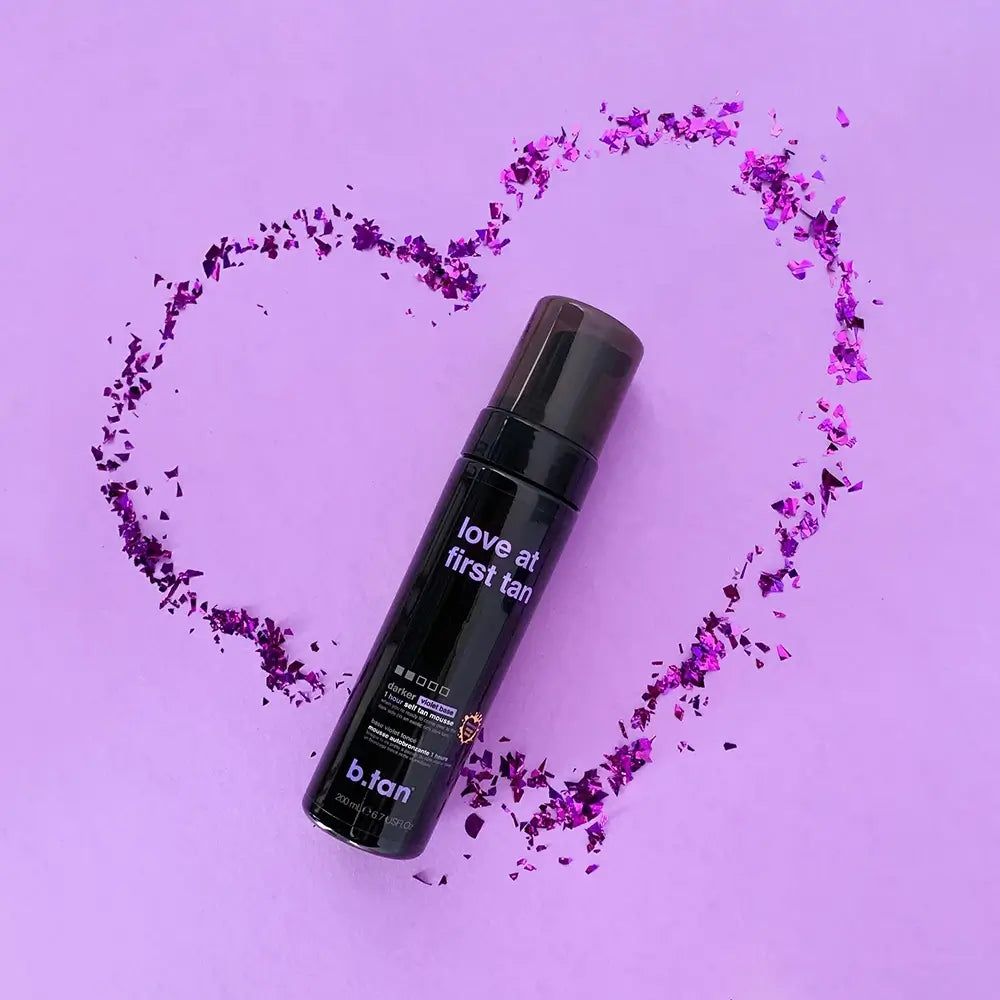 Flacone nero di mousse autoabbronzante circondato da coriandoli viola a forma di cuore. Testo: Love at first tan, b.tan.