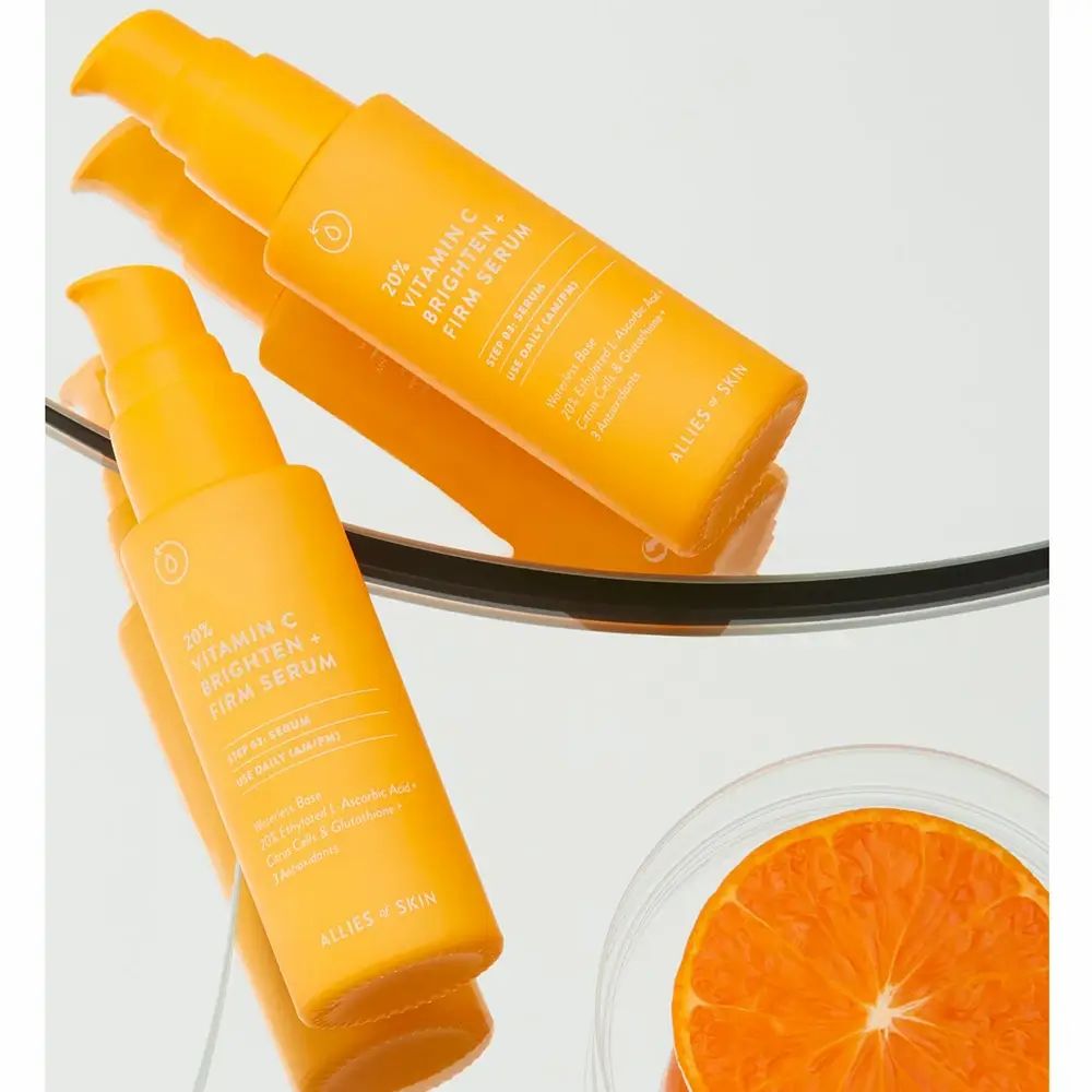 Due flaconi di siero giallo con scritta: 20% Vitamin C Brighten + Firm Serum. Marchio: ALLIES OF SKIN. Su specchio e arancia.