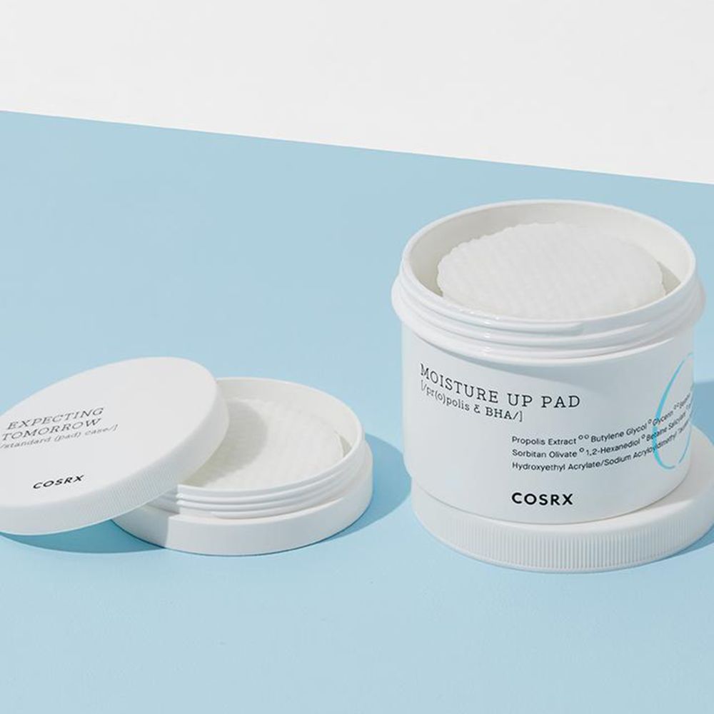 Barattolo bianco aperto con dischetti. Coperchio accanto. Testo: "MOISTURE UP PAD" e "COSRX".