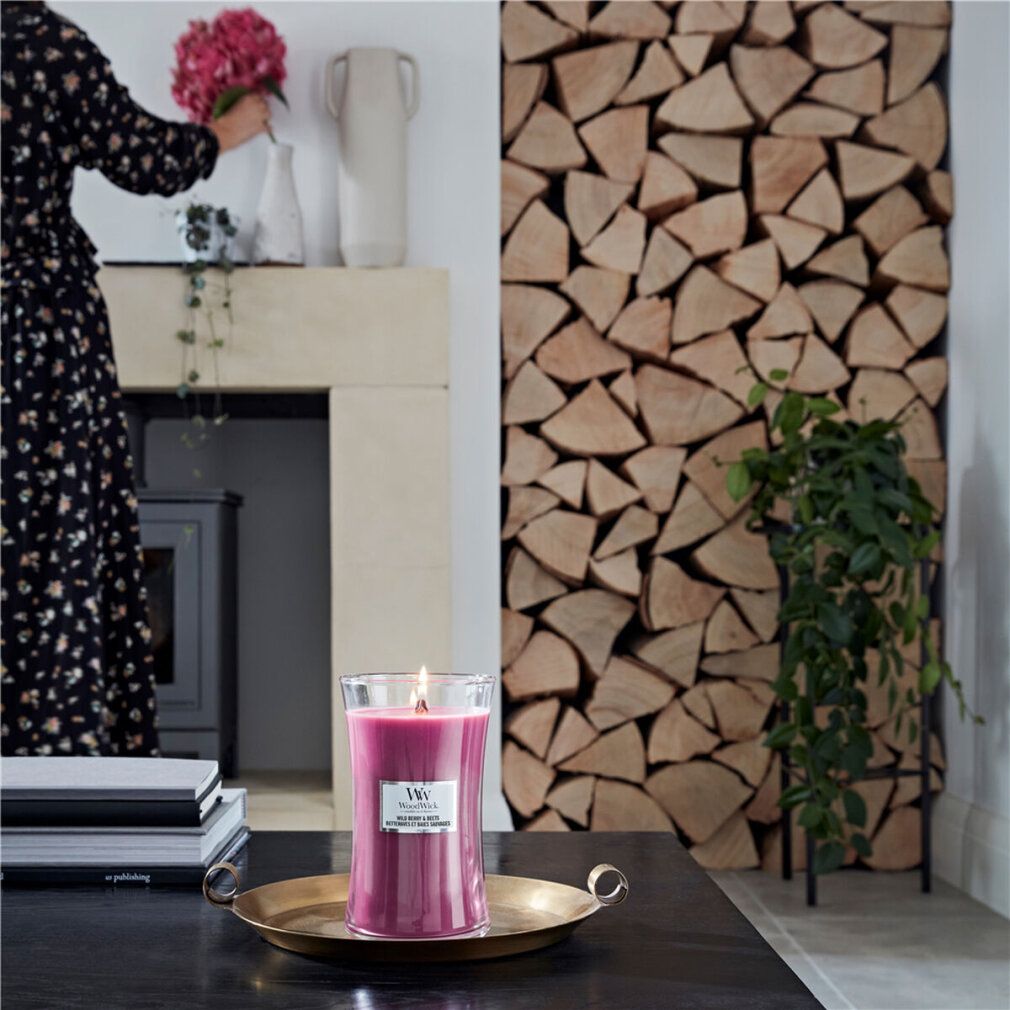 Candela profumata con stoppino in legno accesa su vassoio. Donna con fiori sullo sfondo. Catasta di legna.