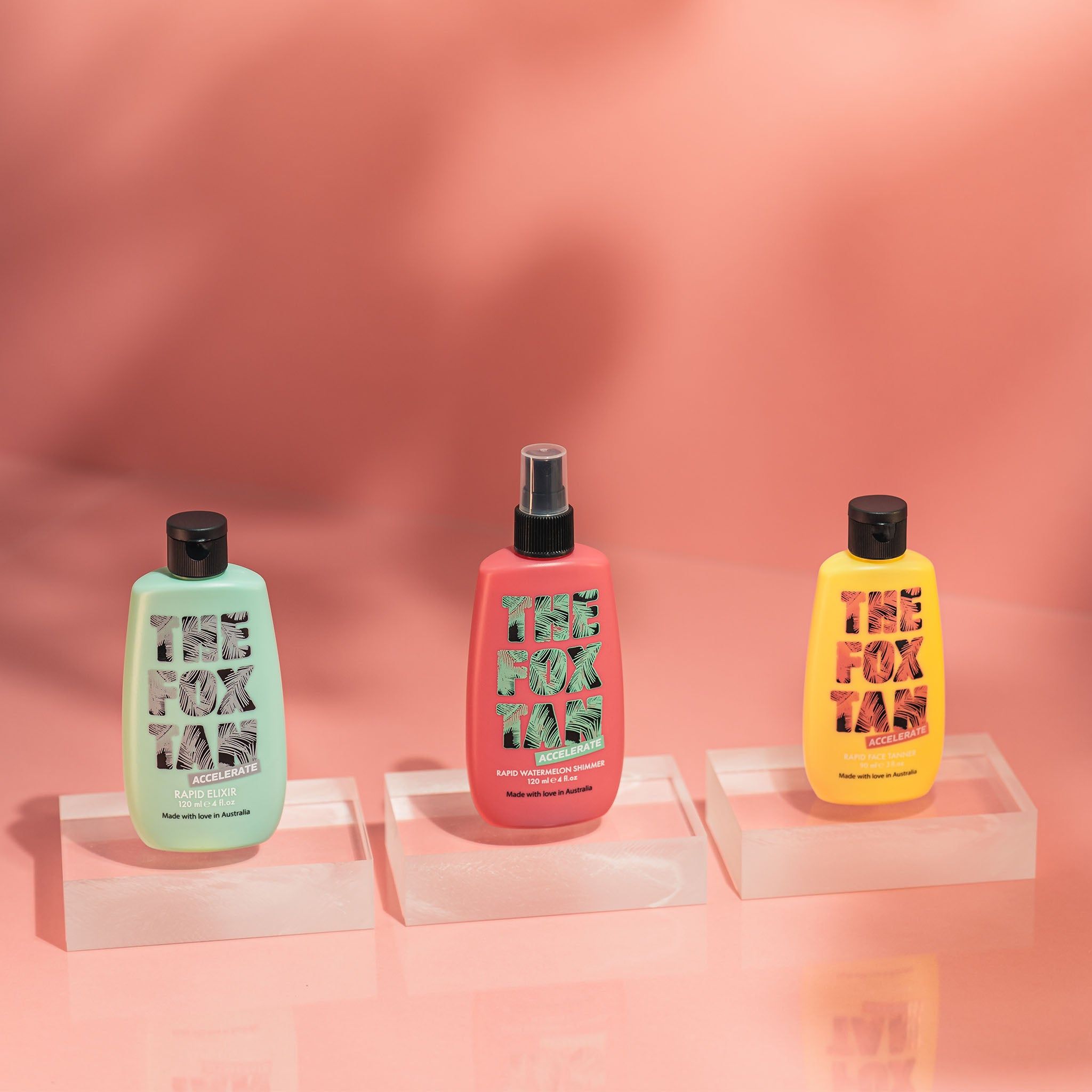 Tre bottiglie su piedistalli. THE FOX TAN. Verde, rosa e giallo. Scritta: ACCELERATE, RAPID ELIXIR. Sfondo rosa.
