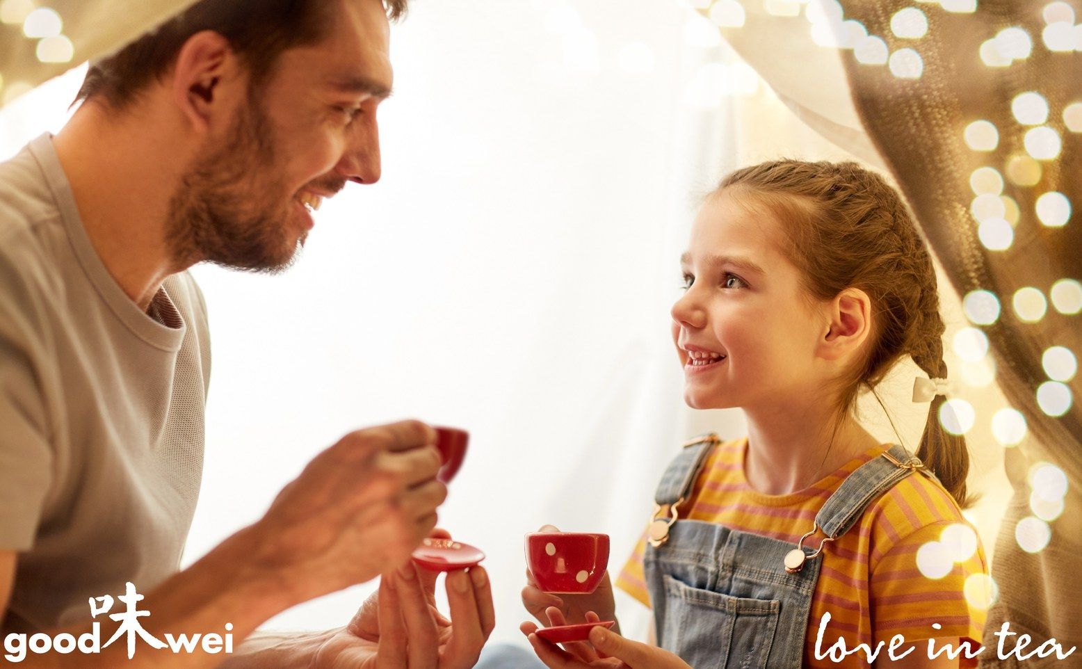 Padre e figlia bevono tè da tazze rosse. Logo "goodwei" e scritta "love in tea" visibili.