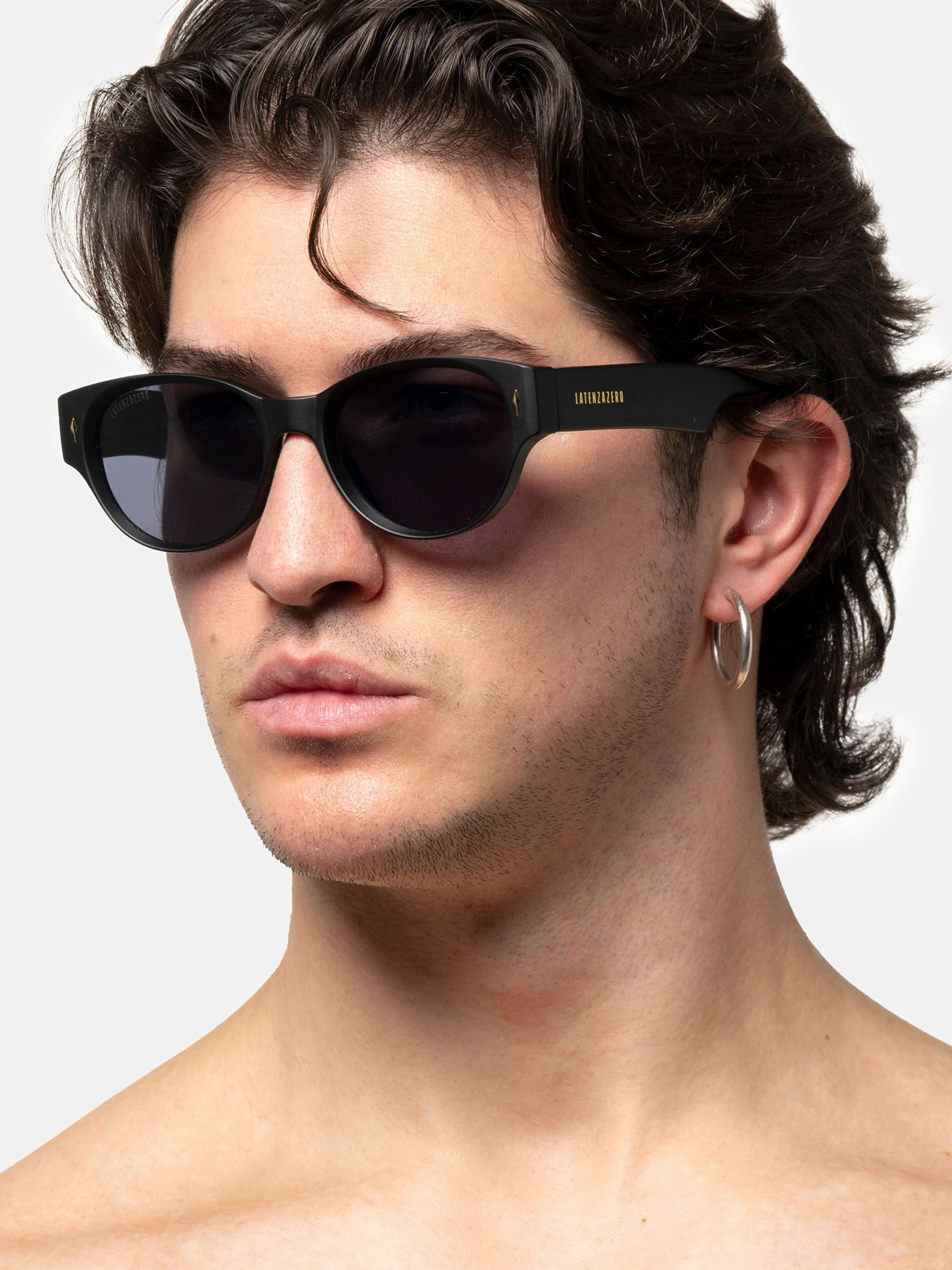 Uomo con occhiali da sole neri. Lenti ovali. Logo Latenzazero. Capelli corti, orecchino.