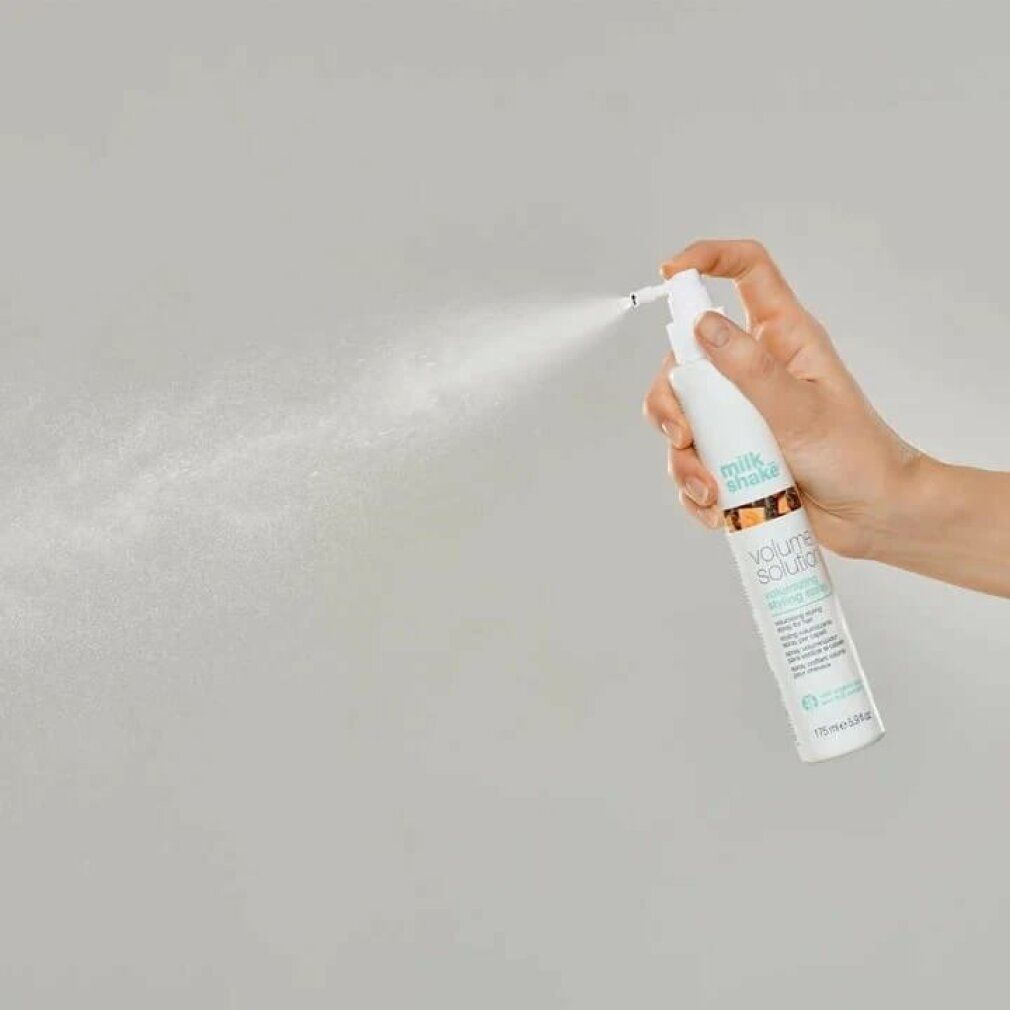 Mano che tiene flacone bianco con erogatore spray. Nebbia spray visibile. Scritta: milk_shake, volume solution, volumizing styling spray.