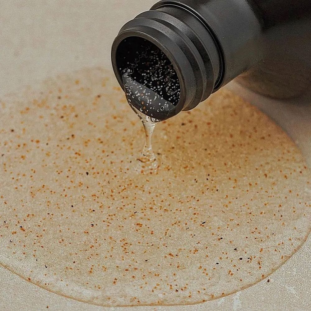 Flacone marrone con erogatore nero. Liquido versato su una superficie. Nessuna indicazione del prodotto visibile.