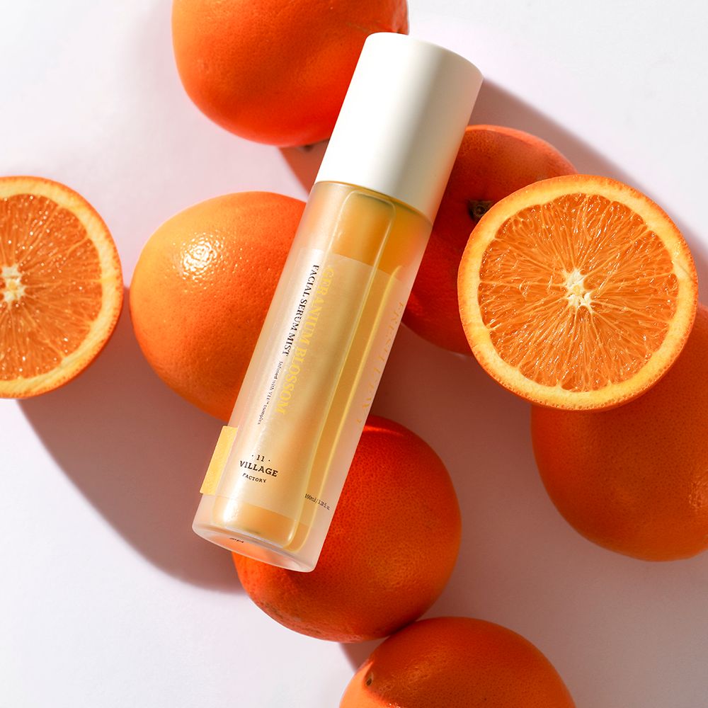 Spray per il viso con tappo bianco, circondato da arance. Etichetta del prodotto visibile. Contenitore trasparente.