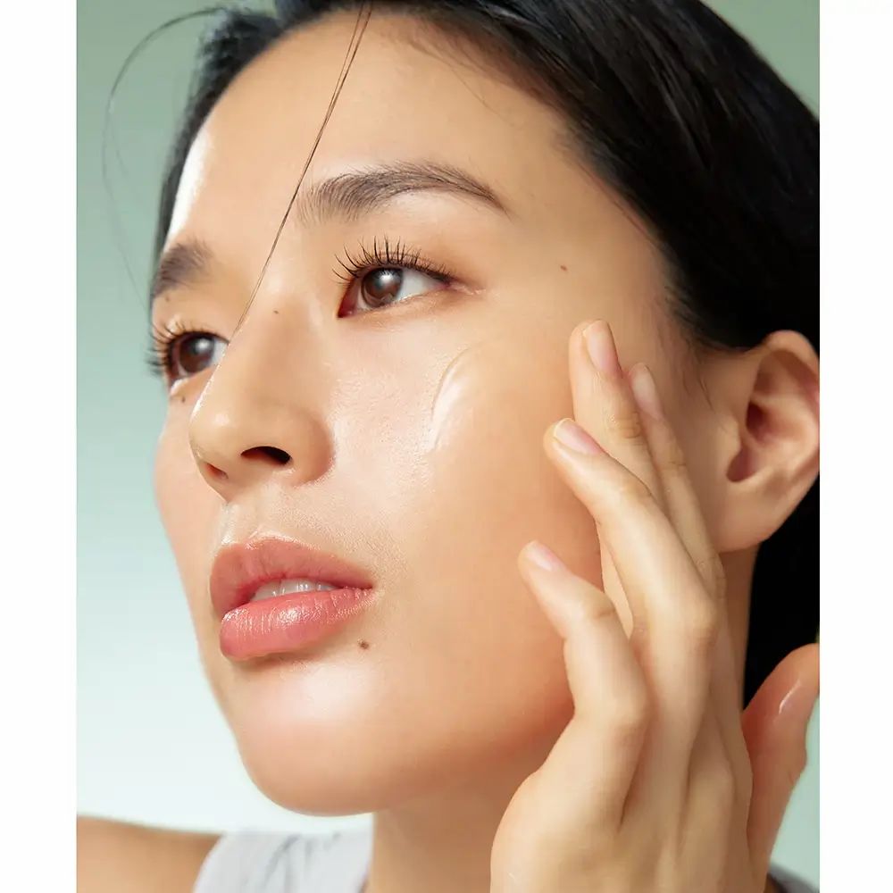 Primo piano di una persona che applica il siero sul viso. Pelle chiara, siero visibile sulla pelle. Ambiente luminoso.