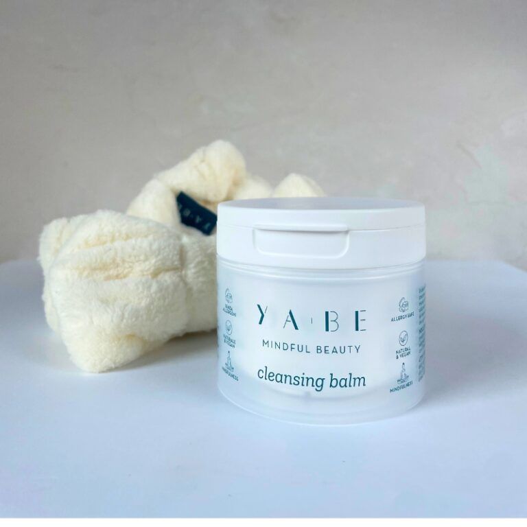 Vasetto bianco con coperchio. Scritta: YA.BE, Mindful Beauty, Cleansing Balm. Accanto a un panno morbido color crema.