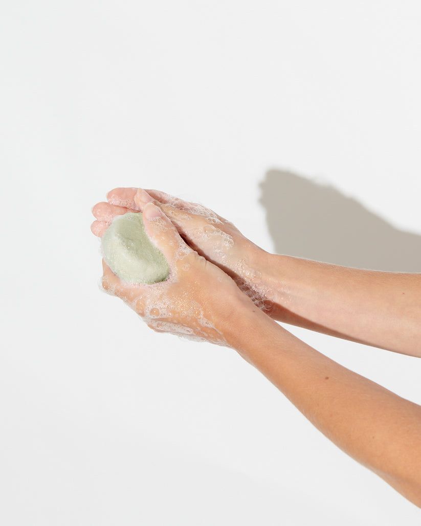 Mani che tengono un panetto shampoo verde rotondo. Schiuma. Su sfondo bianco.