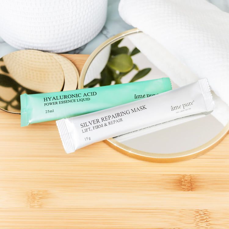 Due bustine di maschera su legno. Verde e argento. Testo: âme pure, Hyaluronic Acid, Silver Repairing Mask. Specchio sullo sfondo.