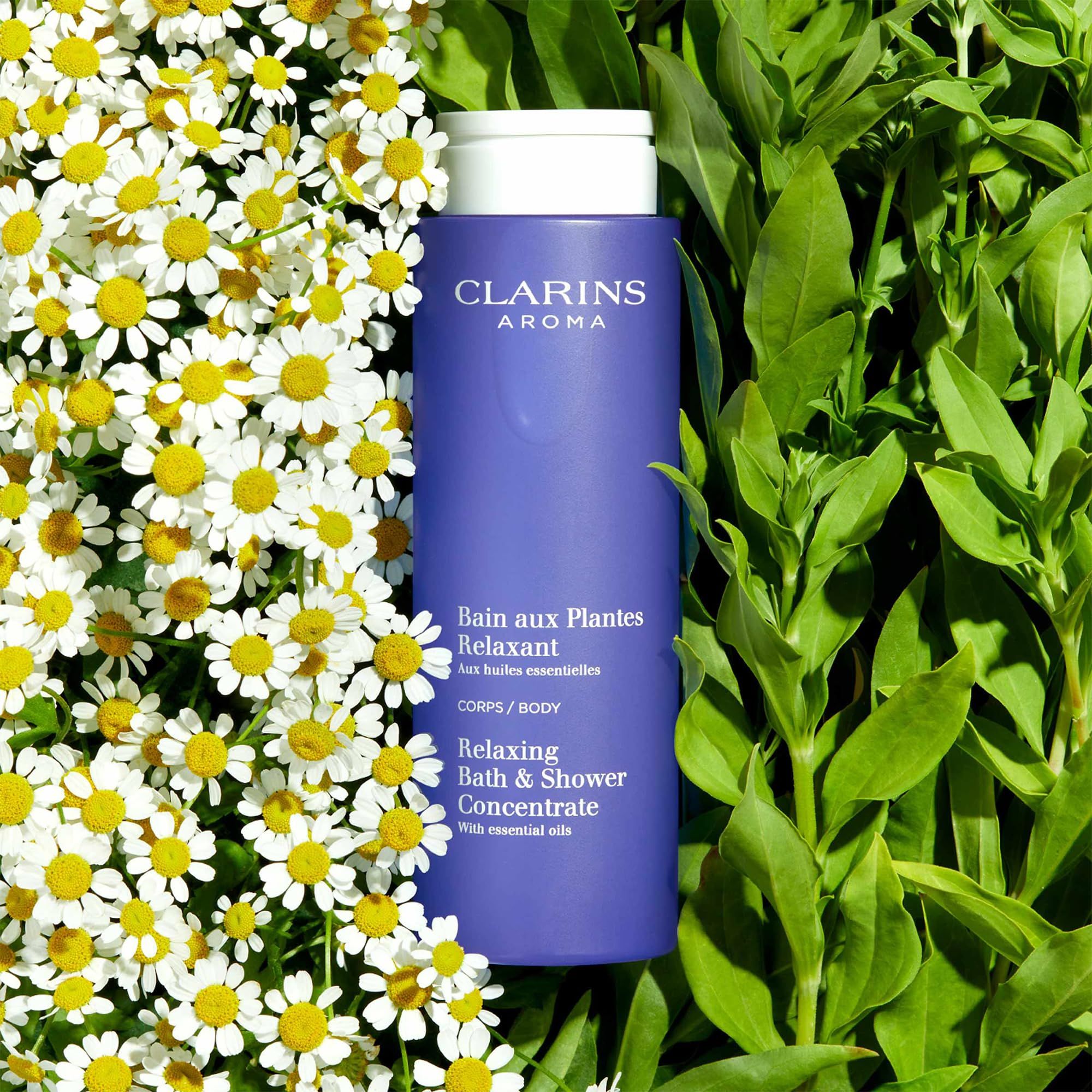 Flacone blu con tappo bianco, circondato da fiori di camomilla e foglie verdi. Testo: Clarins Aroma, Bain aux Plantes Relaxant.