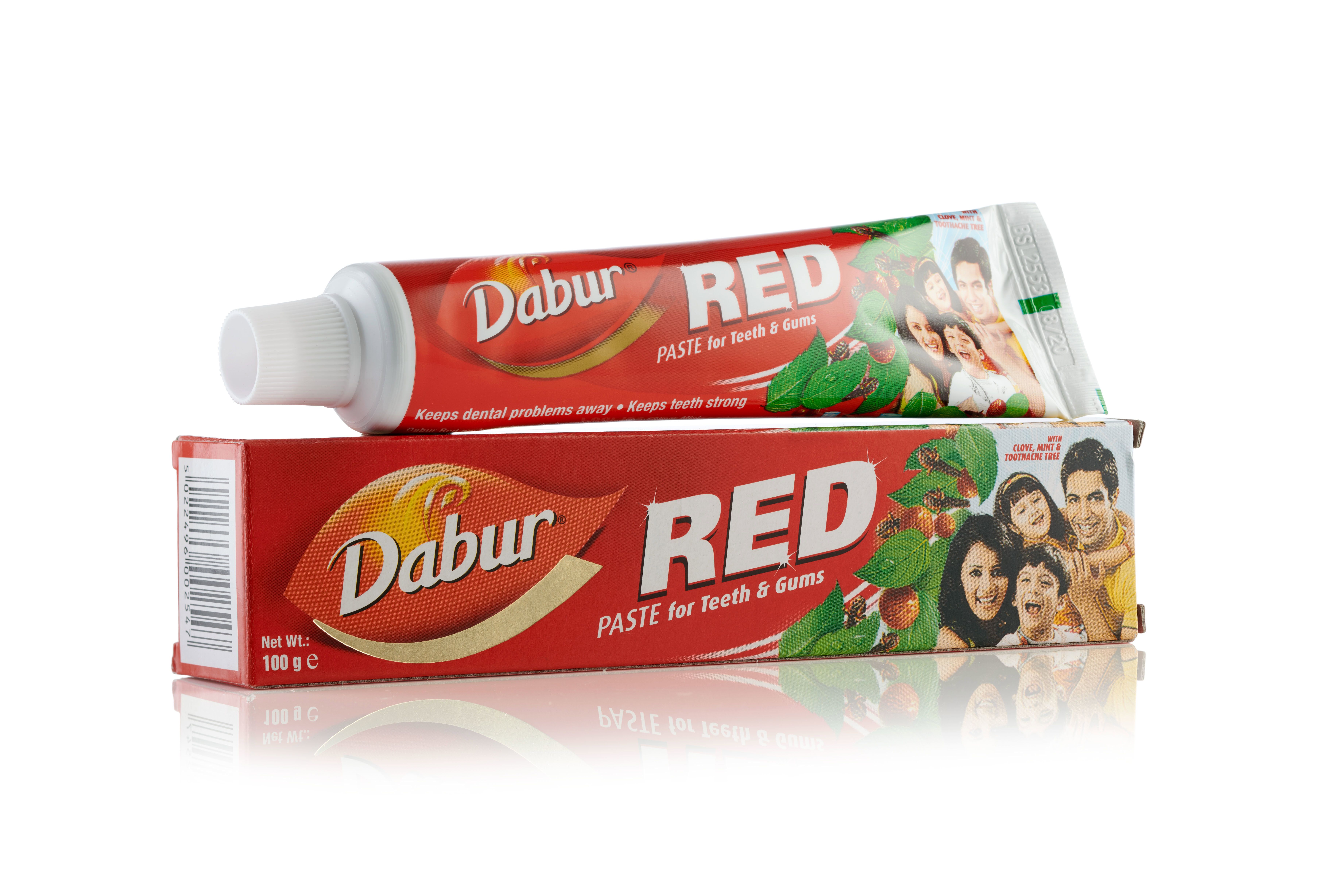 Tubo di dentifricio Dabur Red appoggiato sulla sua confezione rossa. Entrambi mostrano l'immagine di una famiglia.