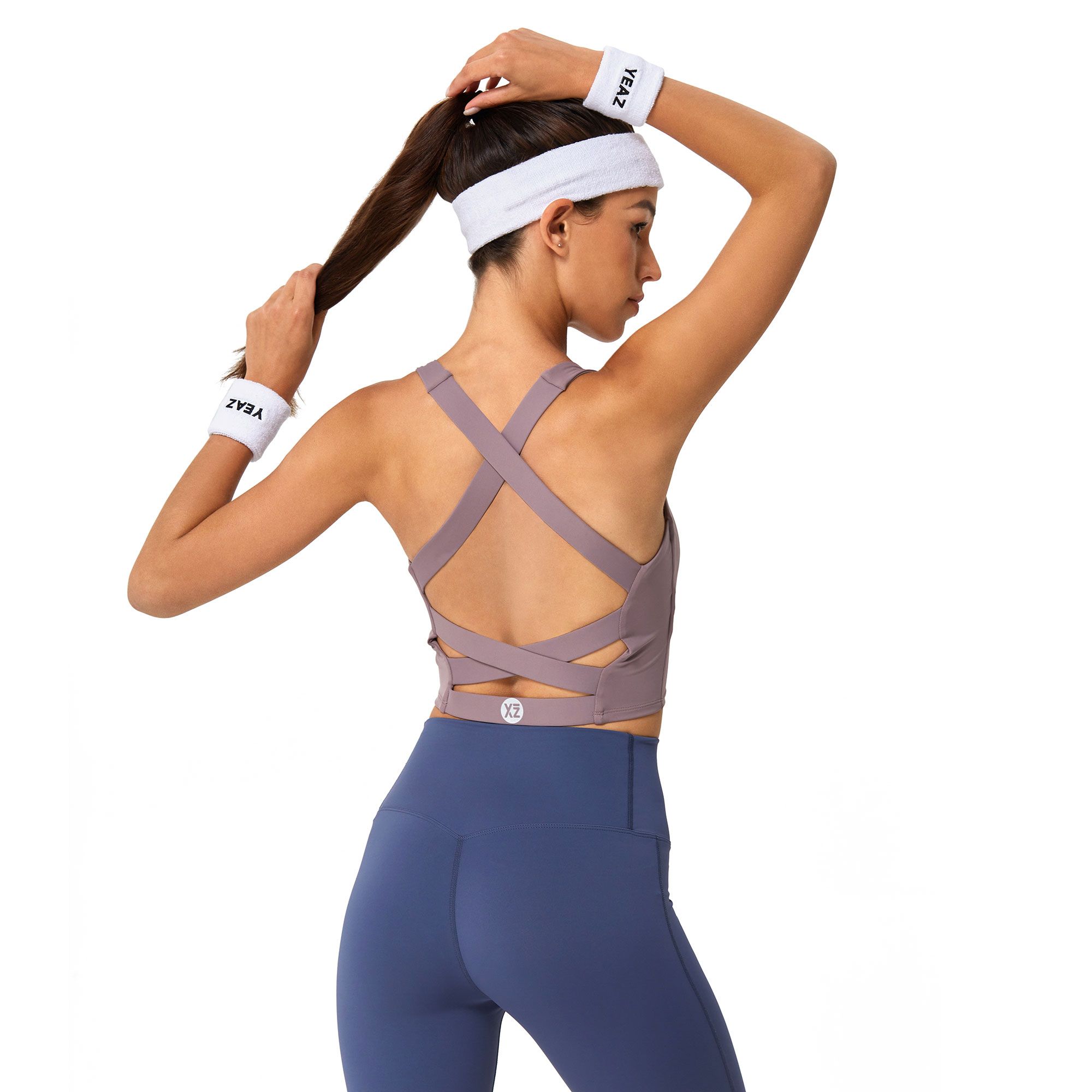 Donna con fasce da polso e fascia per capelli ZYBA indossa un reggiseno sportivo lilla con spalline incrociate e leggings blu.