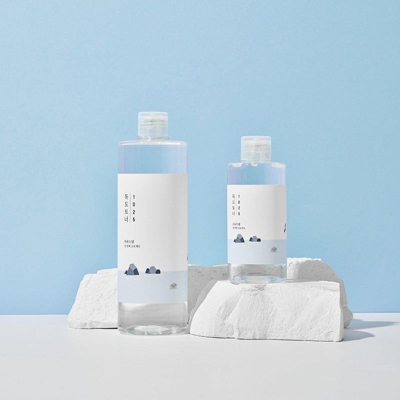 Due bottiglie di liquido trasparente, etichetta bianca e testo coreano. Su piedistallo bianco. Sfondo blu.