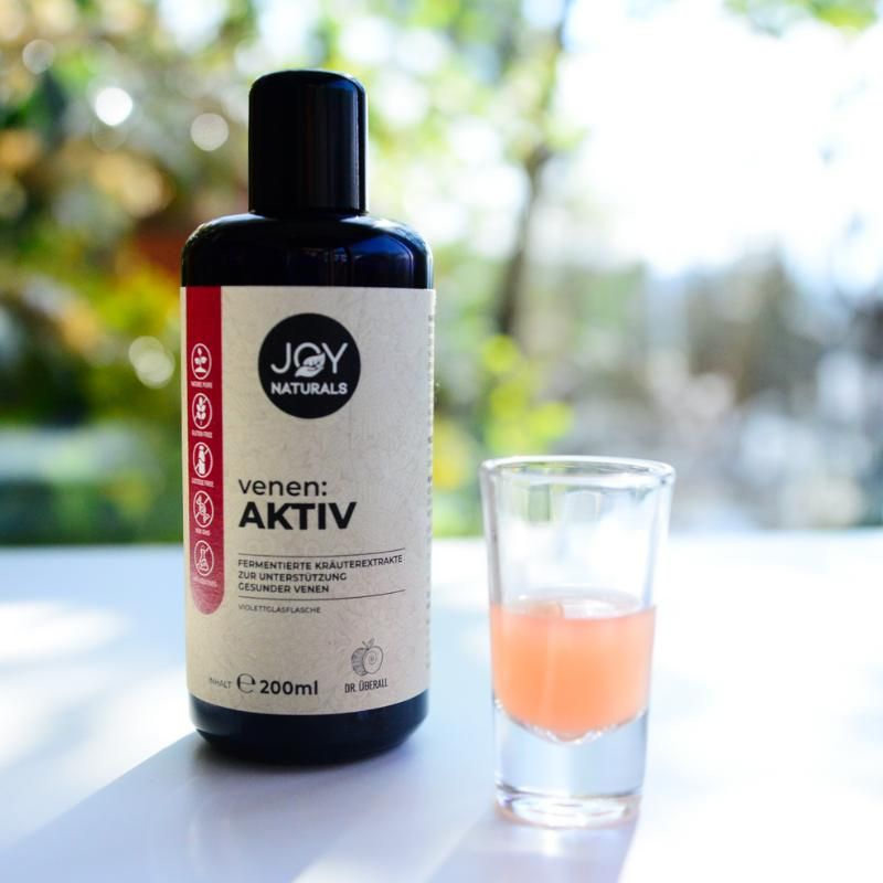 Bottiglia marrone "JOY NATURALS venen:AKTIV" accanto a bicchiere con liquido rosa. Scritte e dettagli sull'etichetta.