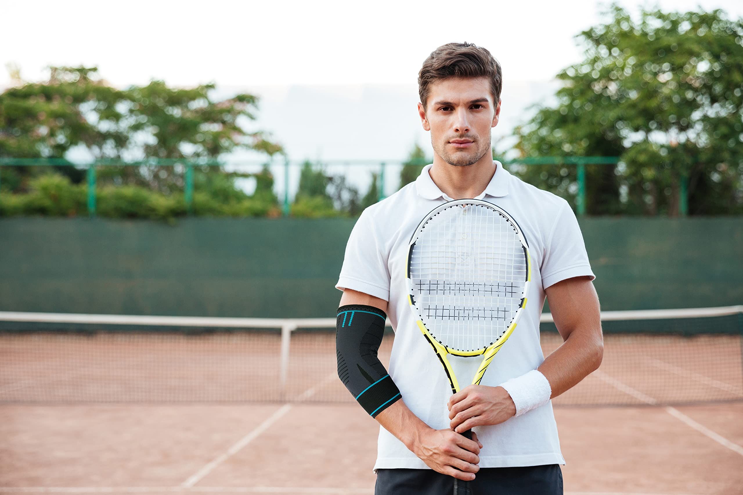 Uomo su un campo da tennis, tiene una racchetta. Indossa abiti bianchi e un tutore per gomito sul braccio sinistro. Rete da tennis sullo sfondo.