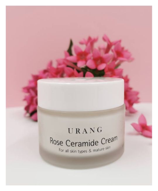 Vasetto di crema con coperchio bianco. Scritta: URANG Rose Ceramide Cream. Per tutti i tipi di pelle e pelli mature. Fiori rosa sullo sfondo.