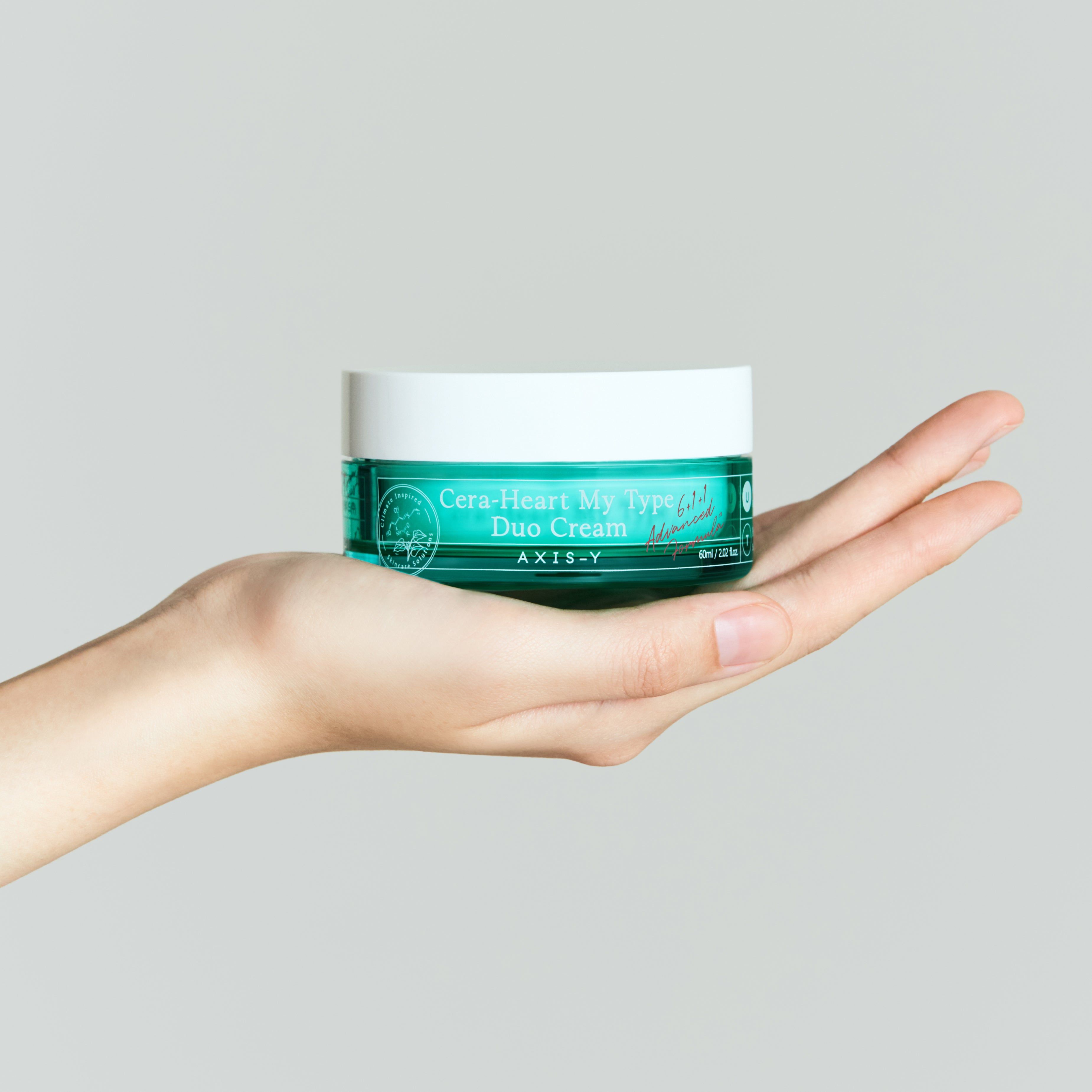 Mano che tiene un vaso di crema verde con coperchio bianco. Scritta: Cera-Heart My Type Duo Cream. Marchio: AXIS-Y.