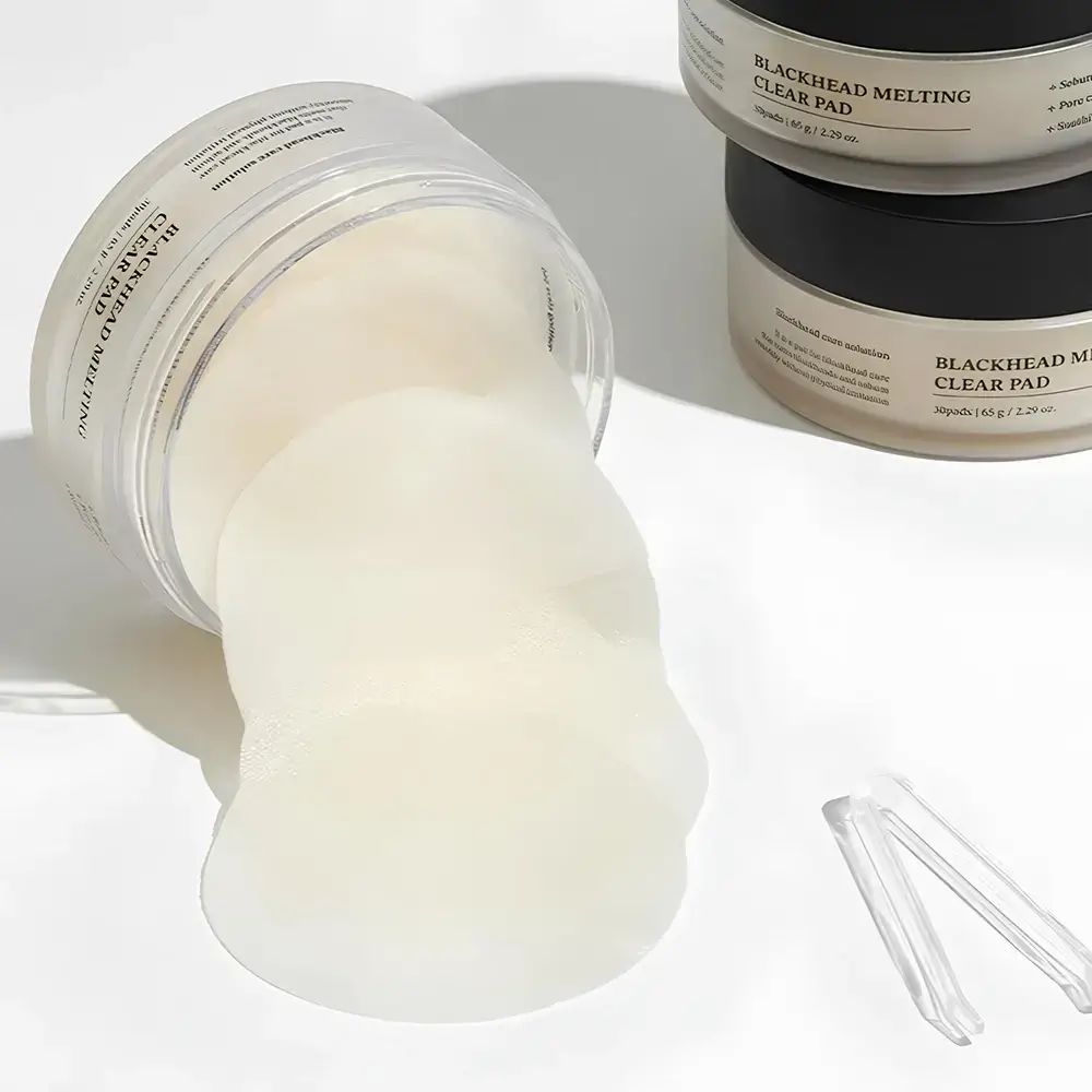 Contenitore aperto con pads. Pinzetta trasparente accanto. Testo: Blackhead Melting Clear Pad.