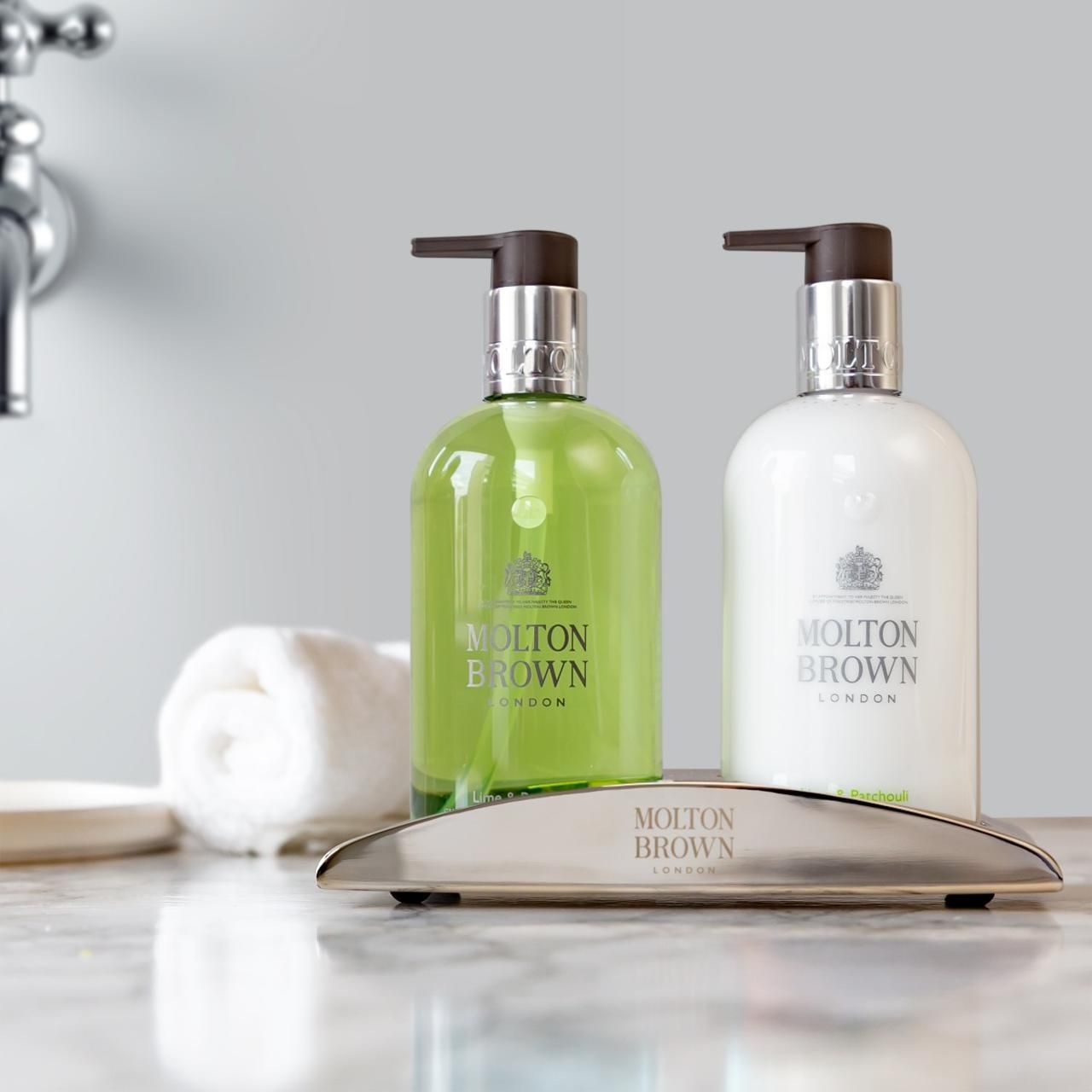 Due flaconi di lozione per le mani con dosatori su un vassoio. Scritta: Molton Brown. Asciugamano e sapone accanto.