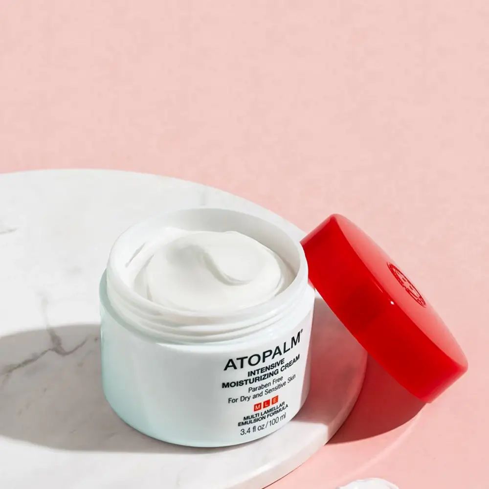 Vaso di crema aperto con coperchio rosso. Scritta: ATOPALM INTENSIVE MOISTURIZING CREAM. Crema su superficie bianca.