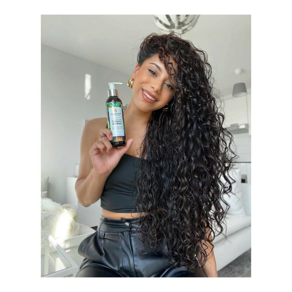 Donna tiene in mano un flacone Protect Me African Citrus Superfruit Hair Oil. Sorride. Lunghi capelli ricci.