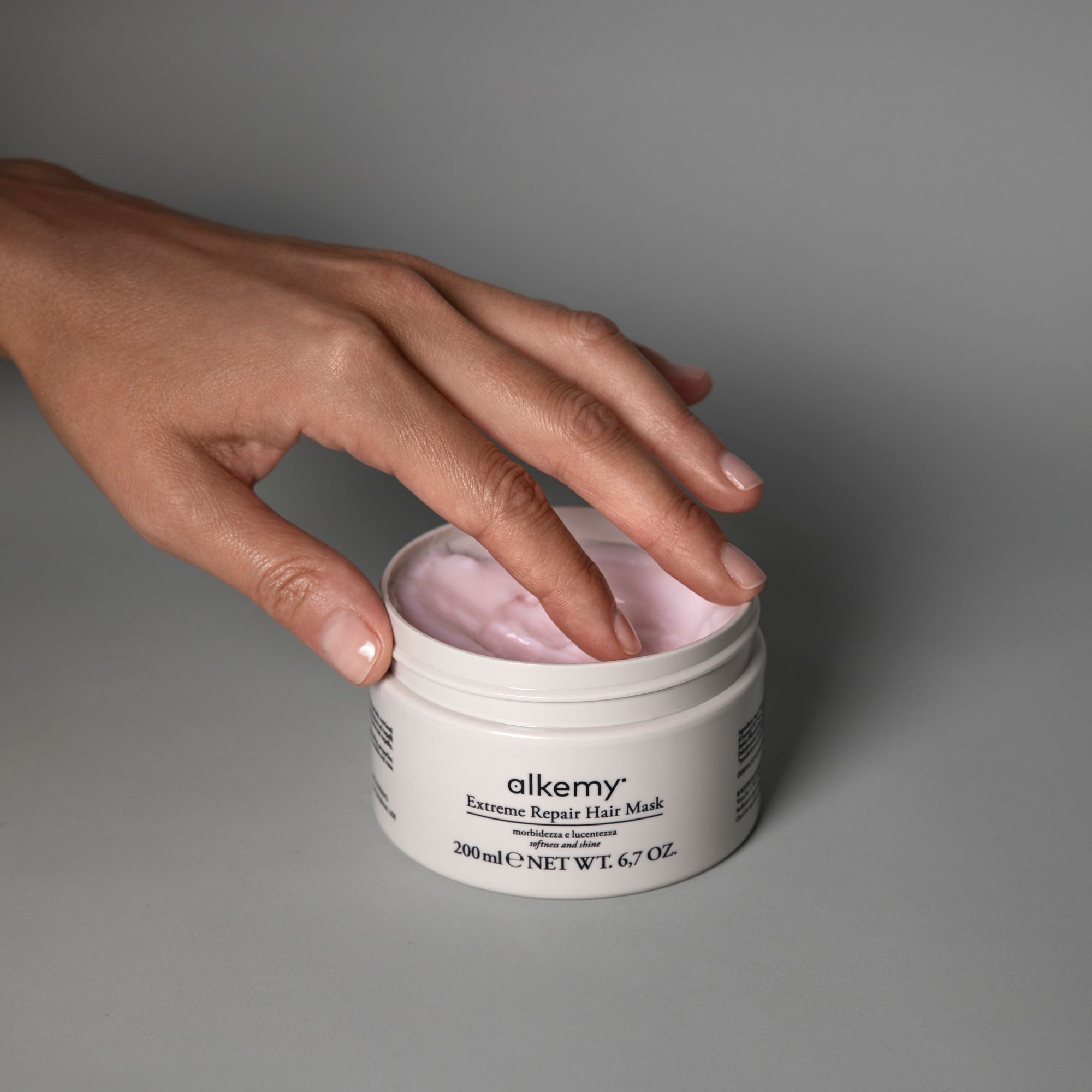 Una mano che affonda in un barattolo aperto di maschera per capelli Alkemy.
