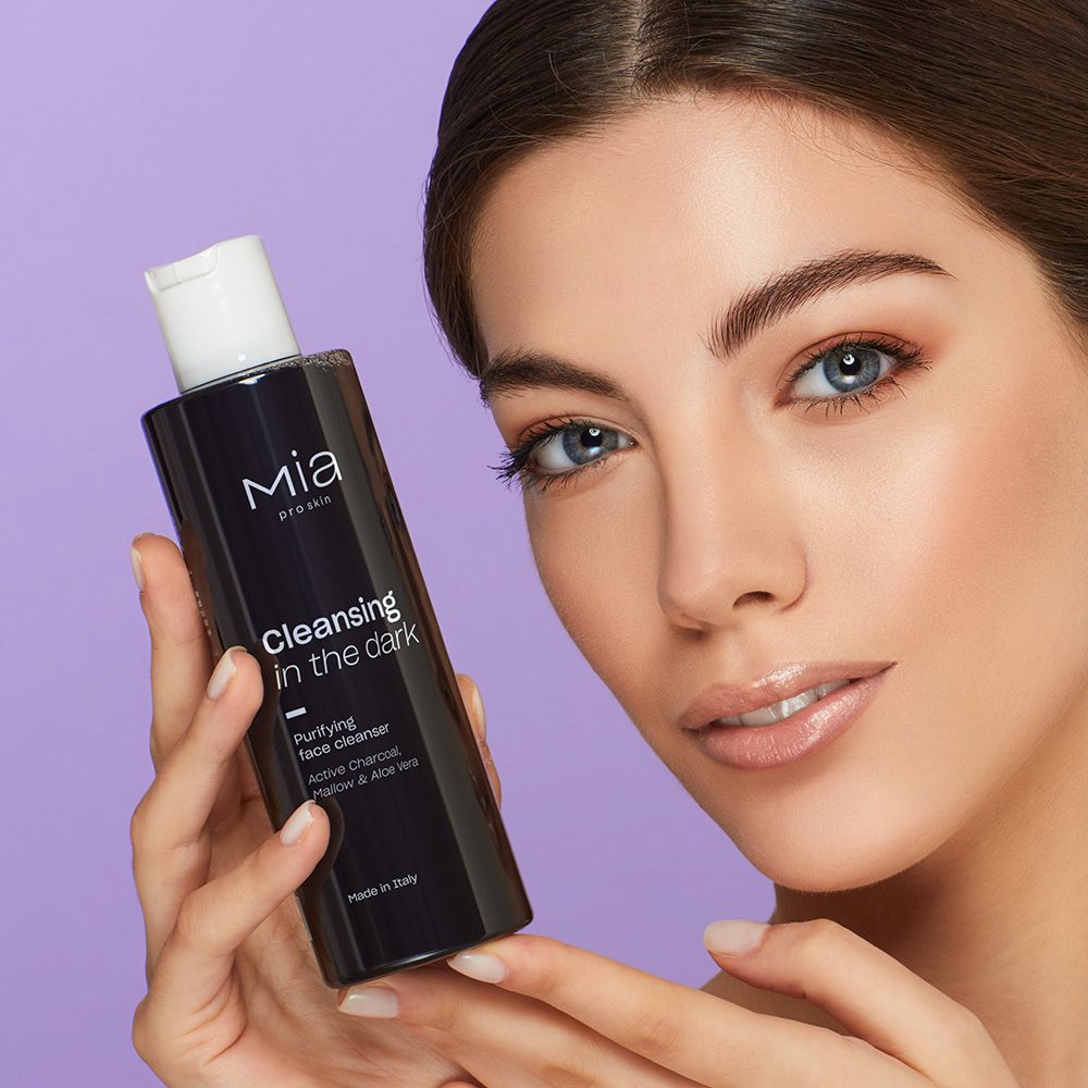 Donna che tiene in mano flacone nero con tappo bianco. Scritta: Mia pro skin, Cleansing in the dark, detergente viso al carbone attivo. Sfondo viola.