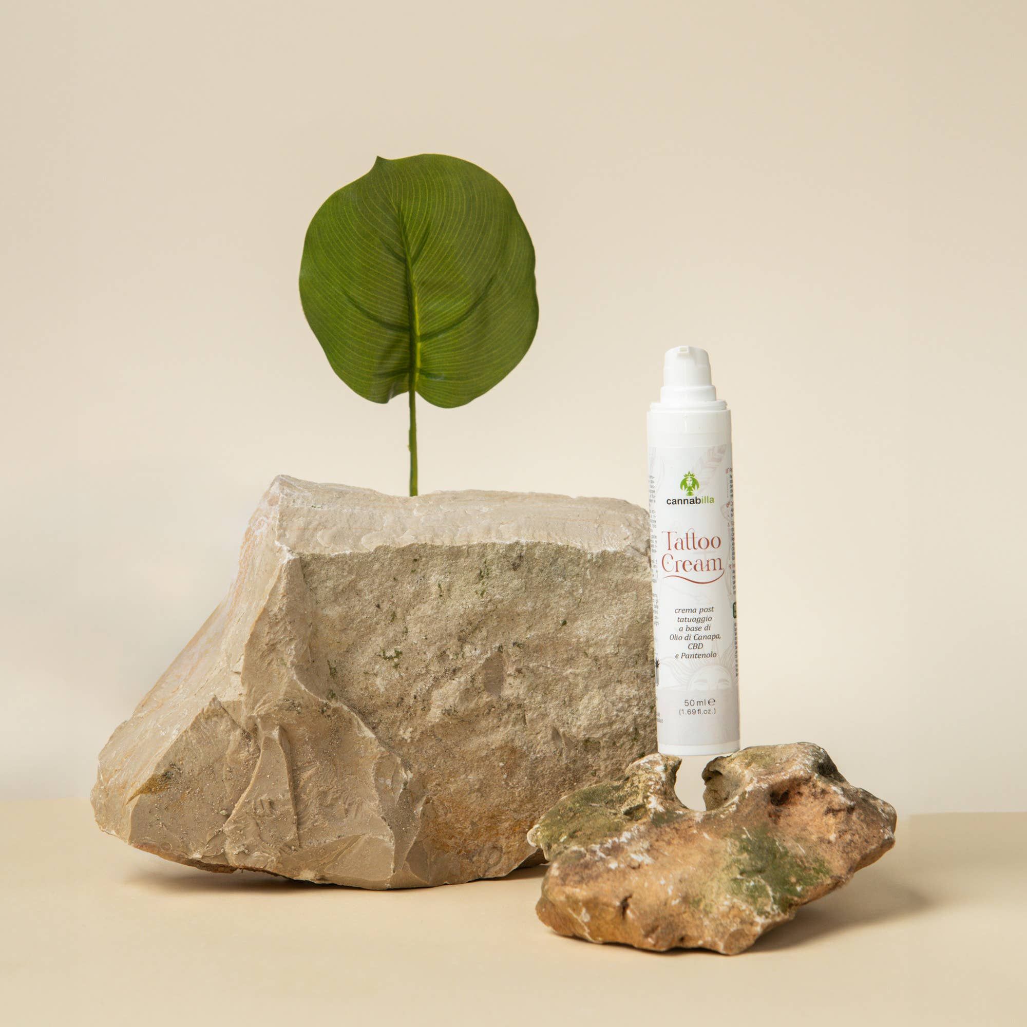 Flacone di Cannabilla Tattoo Cream, posto tra pietre e una foglia. Scritta: Tattoo Cream, crema post tatuaggio, a base di Olio di Canapa, CBD e Pantenolo.