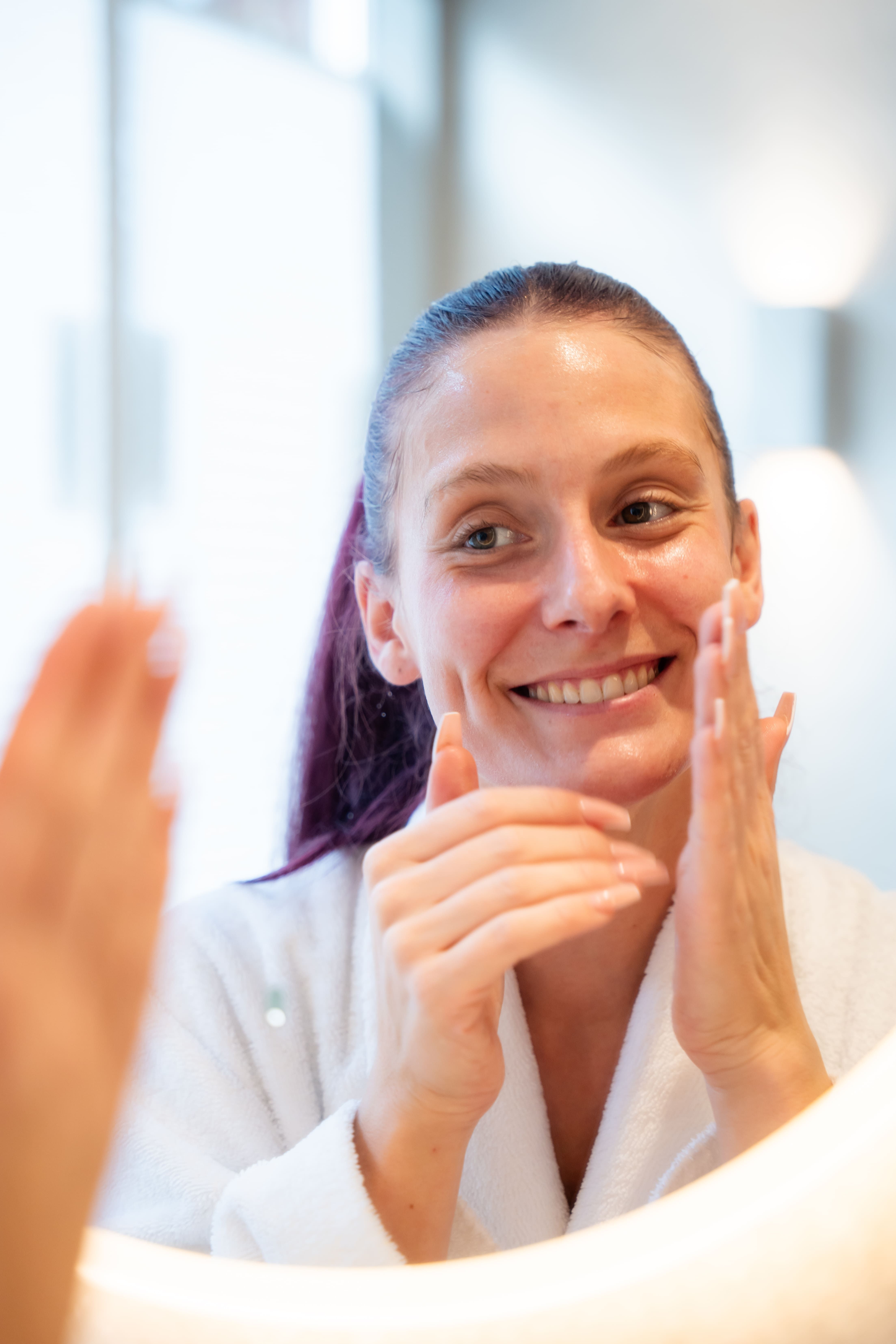 Una donna si applica una crema per la cura della pelle sul viso guardandosi allo specchio.