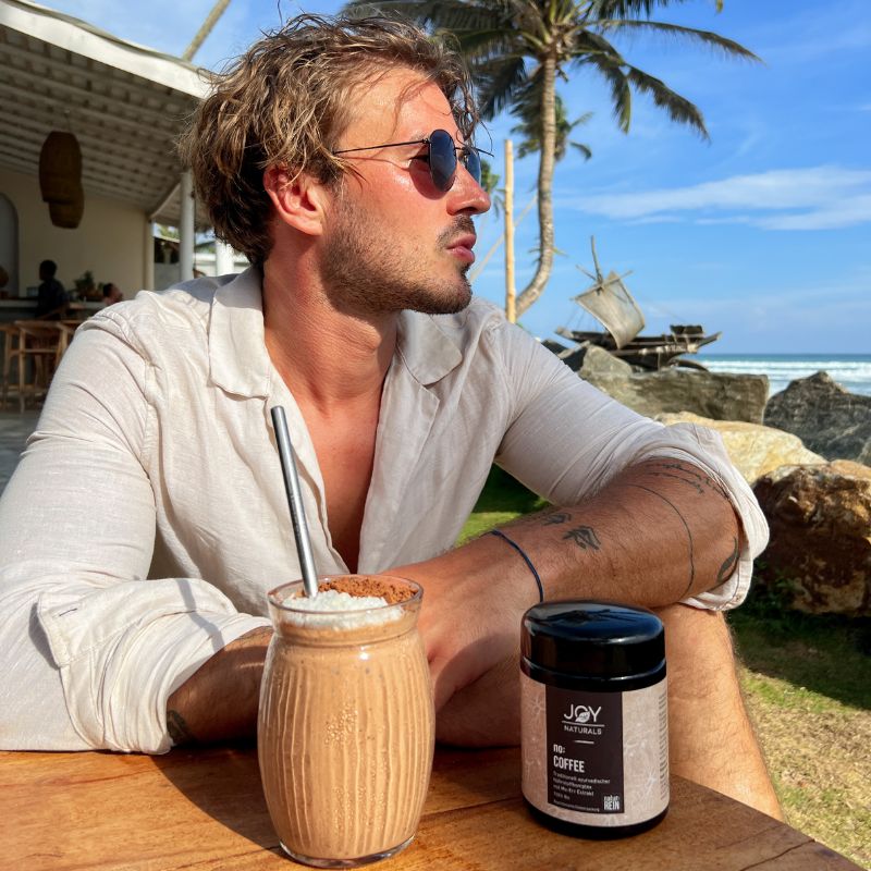 Uomo con occhiali da sole seduto a un tavolo. Accanto, bevanda in vetro con cannuccia e barattolo nero con 'JOY NATURALS no: COFFEE'.