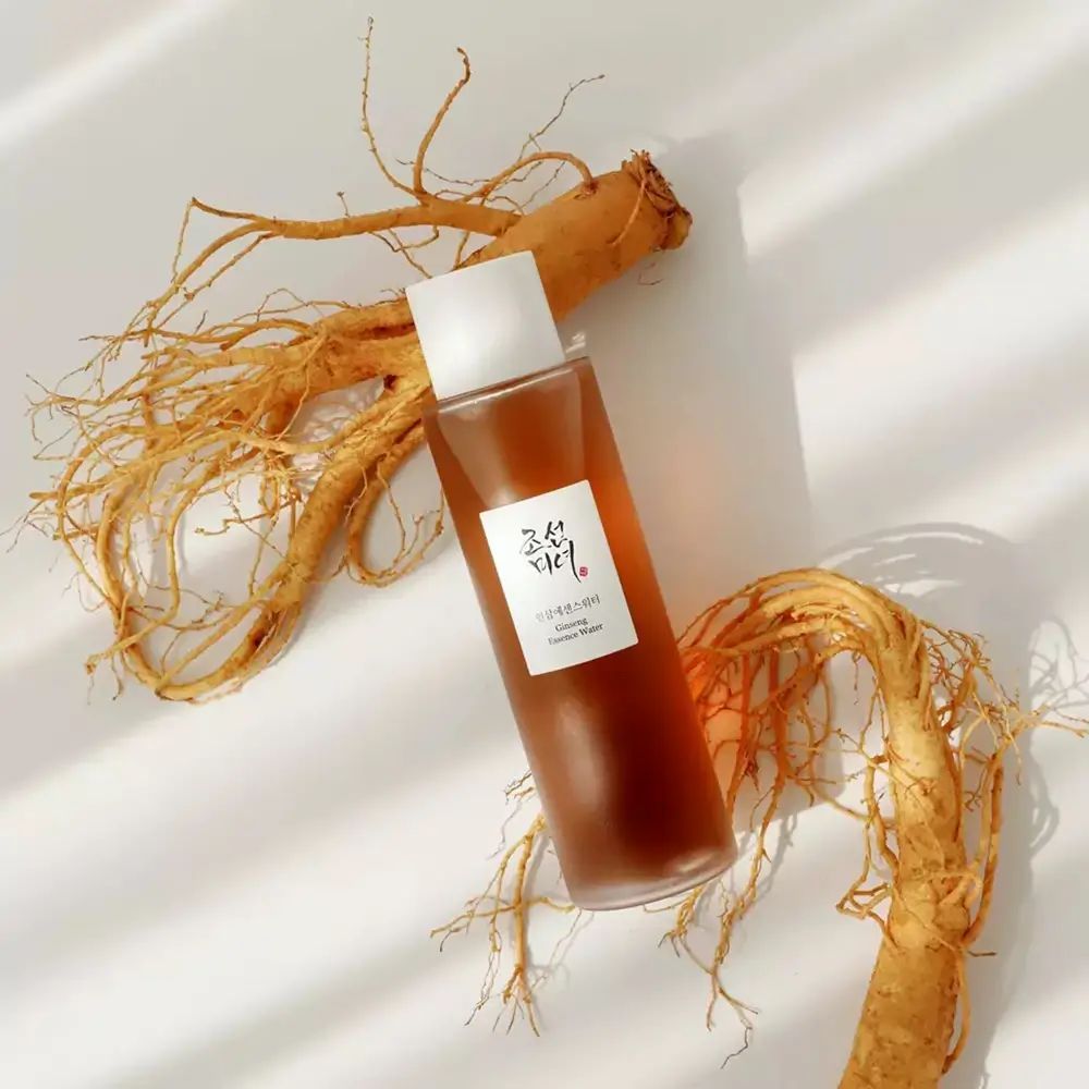 Bottiglia di liquido marrone, circondata da radici di ginseng. Scritta: Ginseng Essence Water. Marchio: Beauty of Joseon.
