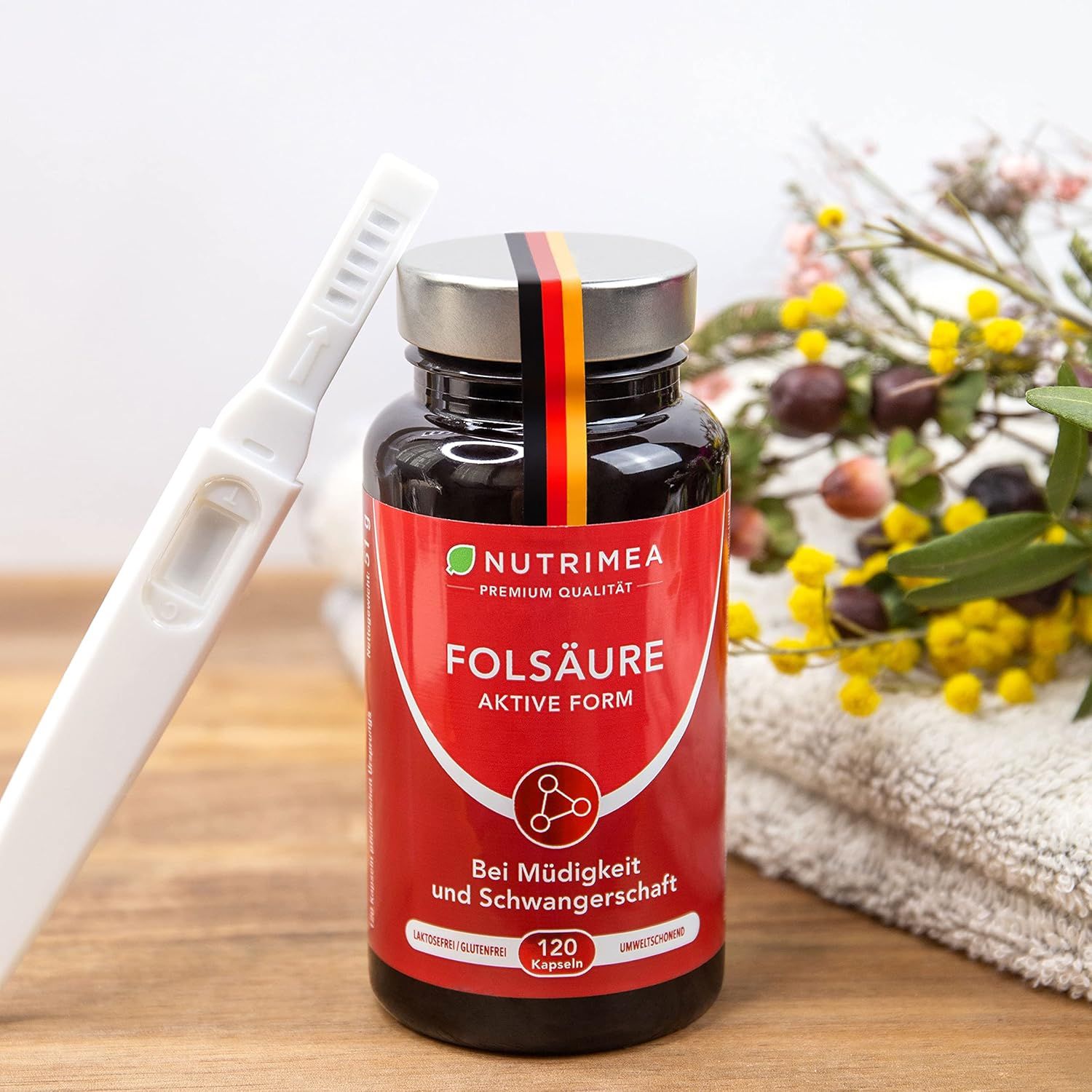 Flacone scuro con etichetta rossa. Contiene capsule di acido folico. Testo: Nutrimea, Folsäure, 120 capsule. Con bandiera tedesca. Test di gravidanza.
