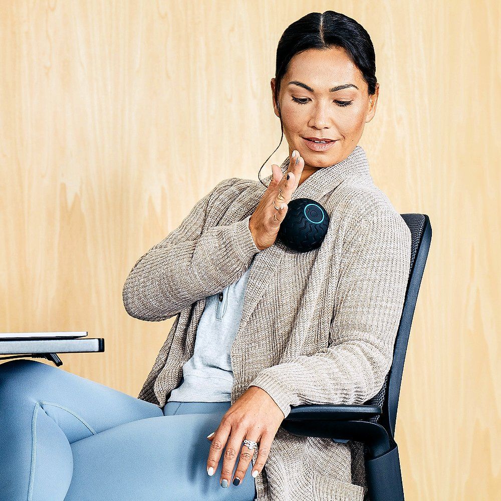 Donna seduta che tiene la sfera da massaggio nera sulla spalla. Indossa un cardigan e pantaloni blu. Sfondo in legno.