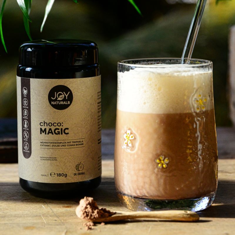 Bicchiere con bevanda e barattolo "JOY NATURALS choco:MAGIC". Cucchiaio con polvere. Bevanda con schiuma. Scritta: Complesso nutritivo con estratto di Triphala, Dulse e fava tonka.