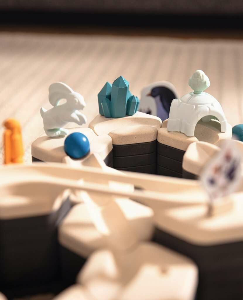 Primo piano di parti del set di gioco GraviTrax Junior. Mostra cristalli blu, un coniglio bianco, una palla blu e una piccola struttura di igloo verde. I pezzi sono disposti su una base grigia. Lo sfondo è sfocato.