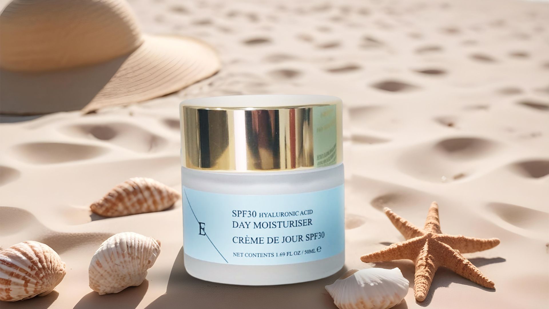 Un vasetto di crema con coperchio dorato ed etichetta blu. Sulla sabbia, con conchiglie, stella marina e cappello sullo sfondo.
